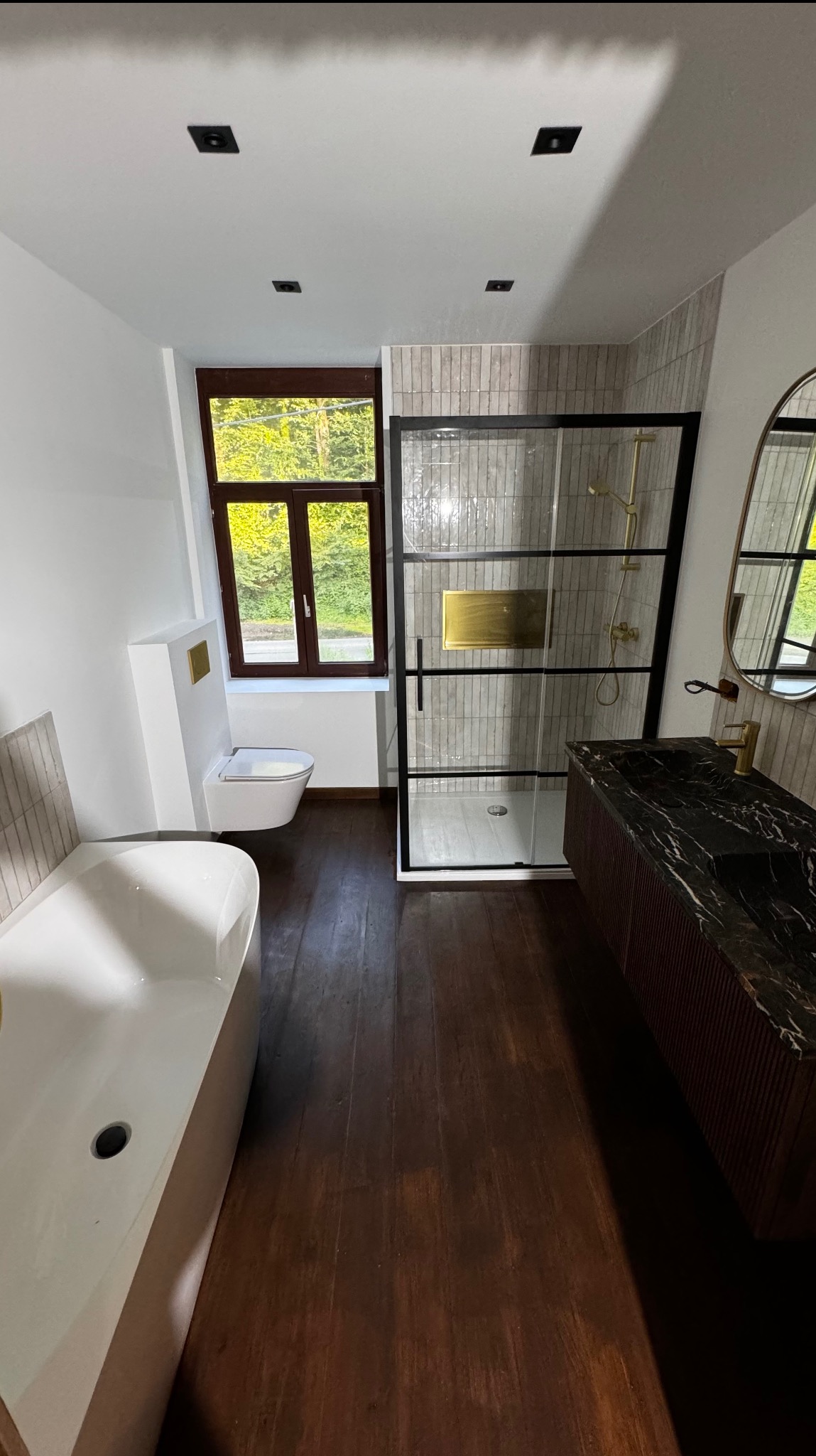 Nowoczesna łazienka z kabiną prysznicową typu walk-in, marmurową umywalką i drewnianą podłogą. Widok na okno z zielenią na zewnątrz. Minimalistyczny design.