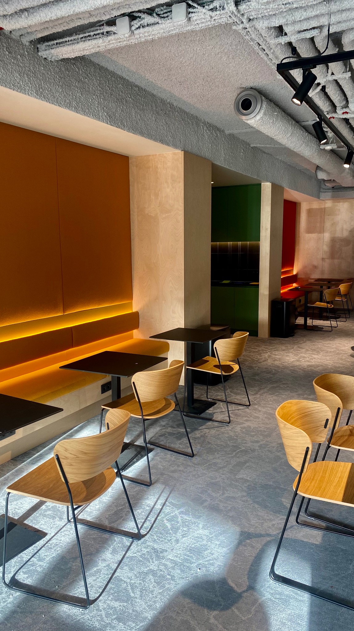 Nowoczesna aranżacja wnętrza biurowego z drewnianymi krzesłami, czarnymi stolikami i oświetleniem LED. Sufit z widoczną instalacją. Kolorowe akcenty w tle.