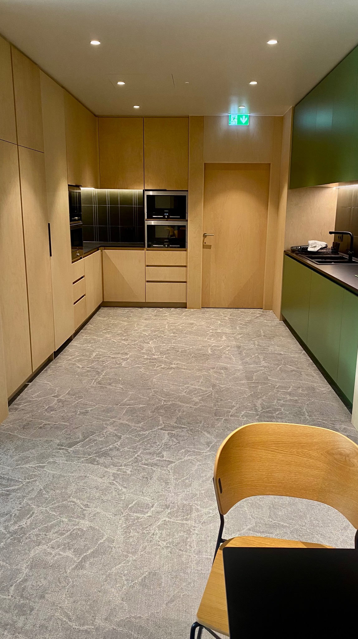Nowoczesna kuchnia z drewnianymi szafkami i szarym marmurkowym dywanem. Zielone akcenty w zabudowie kuchennej. Widoczny fragment krzesła w prawym dolnym rogu.