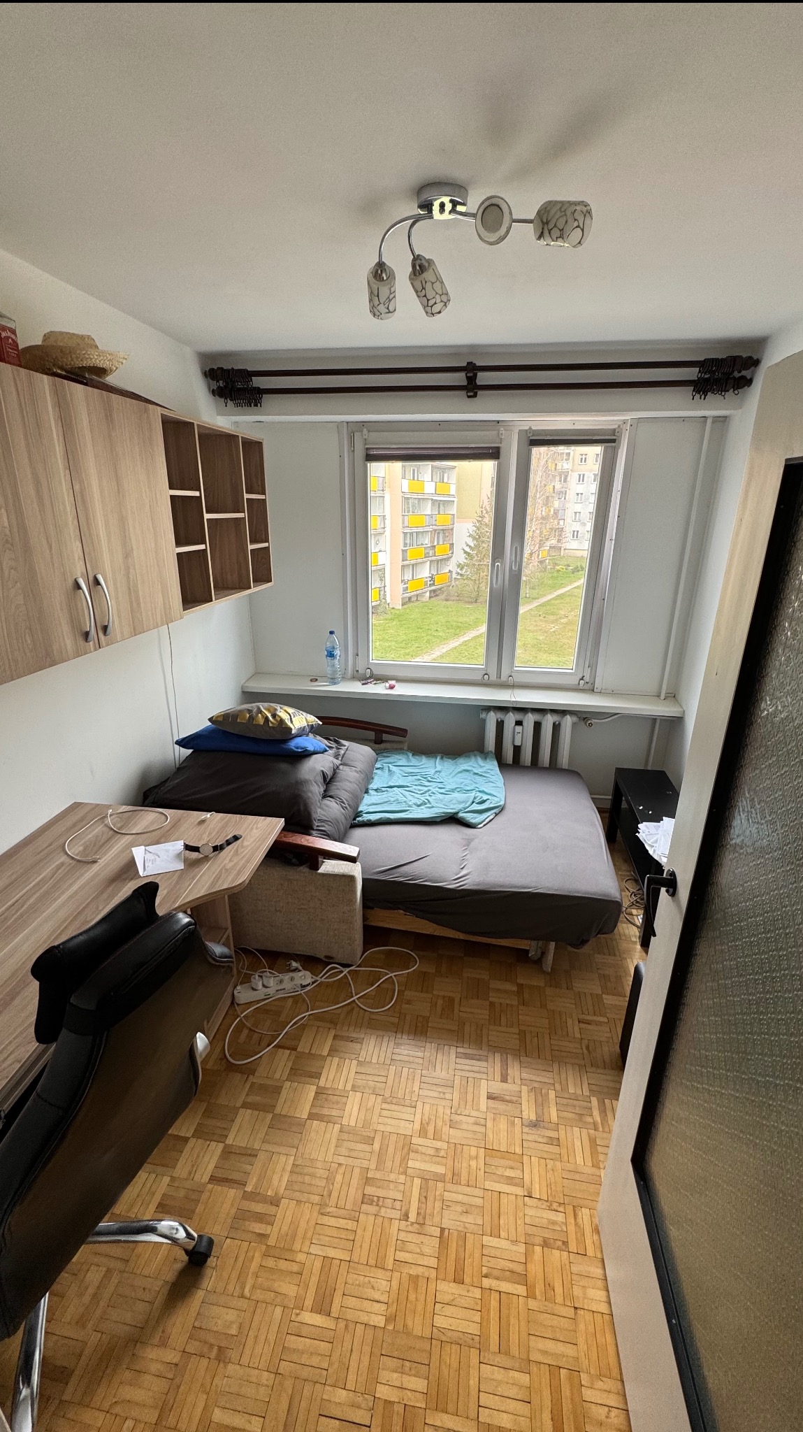 Pokój z parkietem w jodełkę, biurkiem, łóżkiem i oknem z widokiem na blok. Widoczne ślady użytkowania, dodające autentyczności.