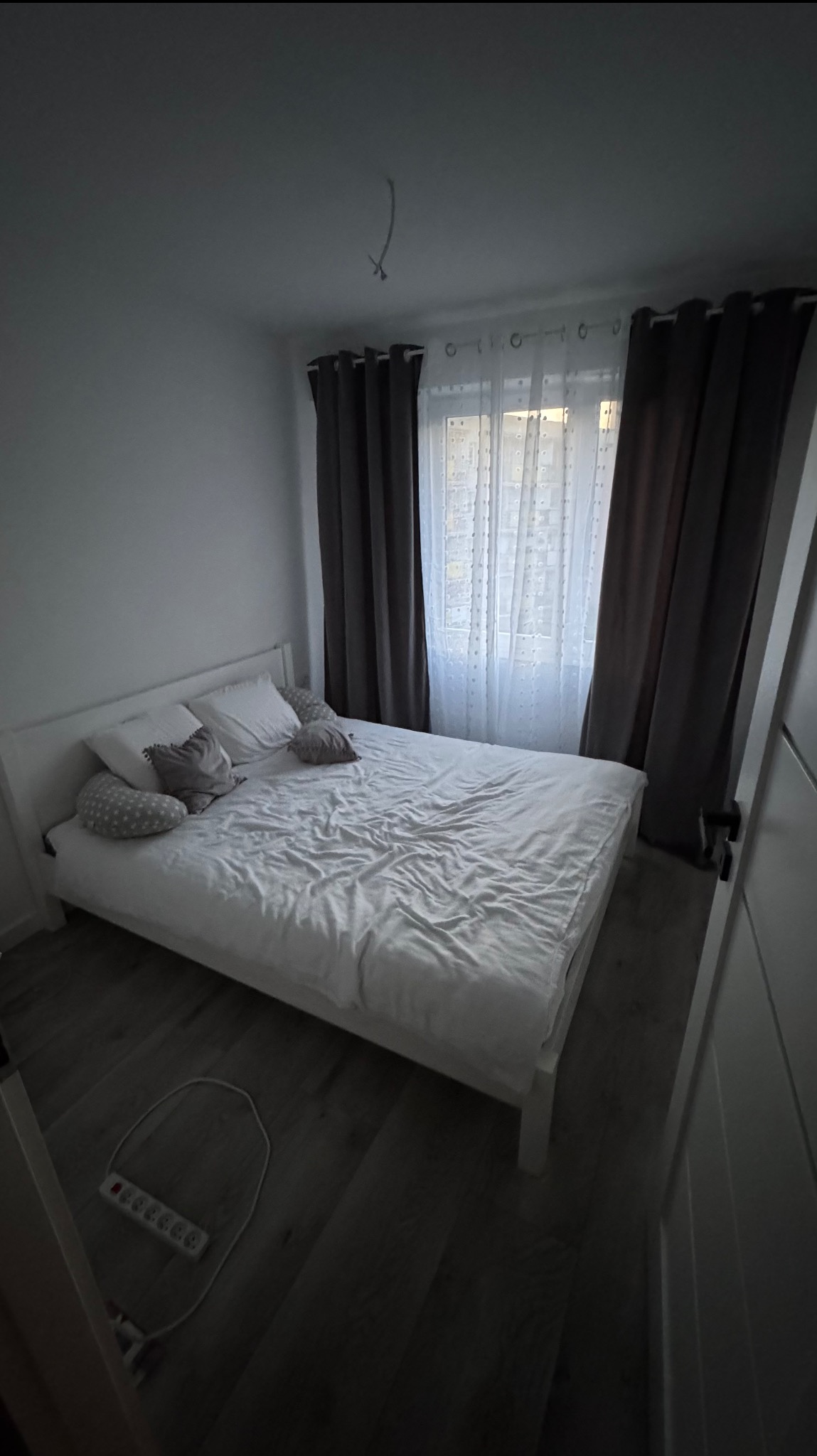 Jasna sypialnia z białym łóżkiem, szarymi zasłonami i drewnianą podłogą. Na podłodze leży przedłużacz. Widok na okno z delikatną firanką.