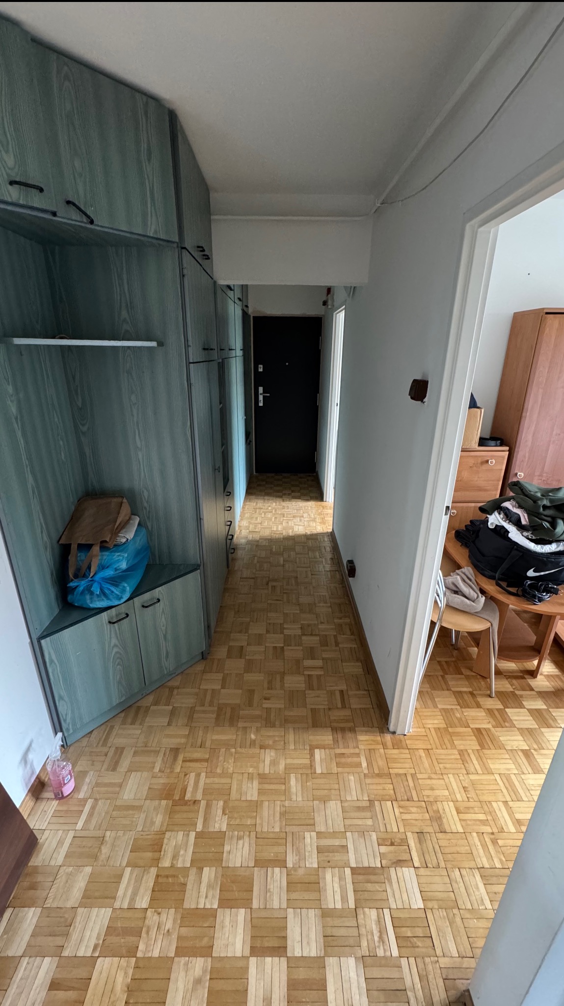 Korytarz z parkietem w kwadraty, zabudowa szaf w odcieniach szarości. Widok na drzwi wejściowe i otwarte przejście do innego pomieszczenia. Perspektywa z głębi mieszkania.