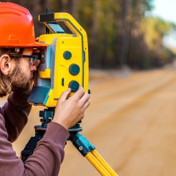 Geodeta - Usługi geodezyjne - Pomiar gruntu: mężczyzna w kasku i kamizelce, obsługuje żółty teodolit na trójnogu, w tle droga gruntowa i drzewa. Precyzyjne pomiary w terenie.