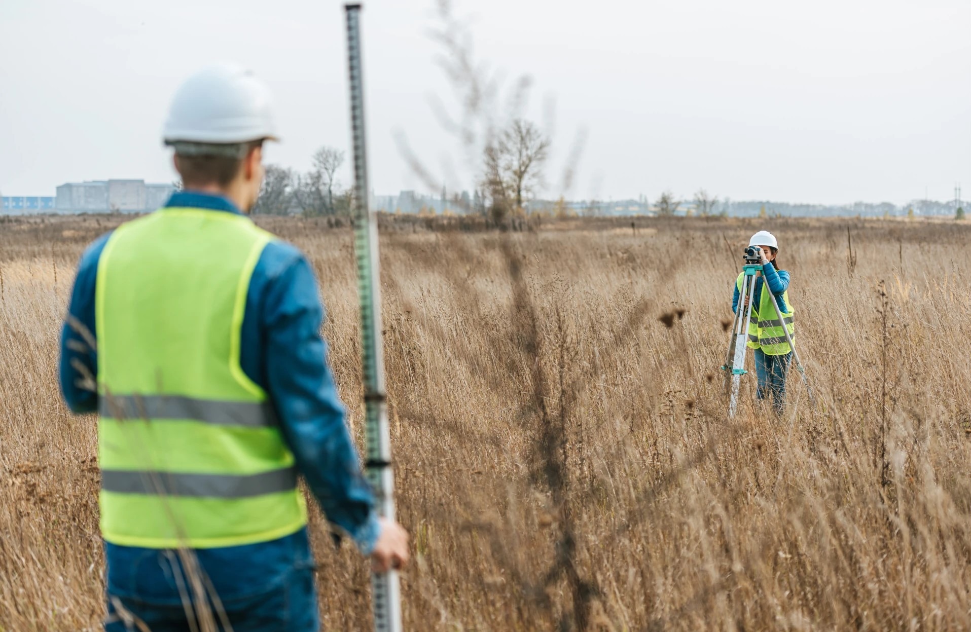 Dwóch pracowników w kamizelkach i kaskach, wykonujących pomiary geodezyjne na zewnątrz, na tle pola z wysoką trawą i budynków w oddali.