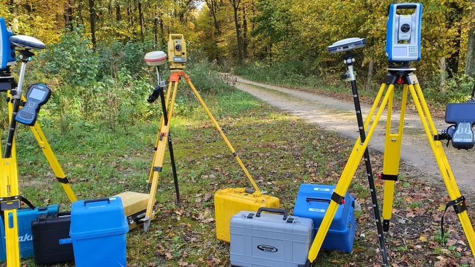 Sprzęt pomiarowy na żółtych statywach w terenie leśnym. Widoczne skrzynie transportowe. Urządzenia gotowe do pracy.