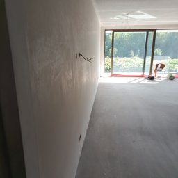 Usługi budowlane Piotr Skraburski - Ściana po szpachlowaniu z wystającymi kablami elektrycznymi. W tle widoczne okno balkonowe i osoba pracująca w jasnym, przestronnym wnętrzu.