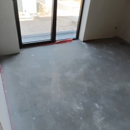 Usługi budowlane Piotr Skraburski - Wnętrze pokoju z wylewką samopoziomującą, przygotowane do dalszych prac wykończeniowych. Widoczne duże okno z czarną ramą i fragment budynku na zewnątrz.