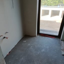 Usługi budowlane Piotr Skraburski - Surowe wnętrze w trakcie remontu: szpachlowane ściany, wylewka podłogowa, wystające kable i rury, okno z widokiem na zieleń. Etap przygotowania do dalszych prac wykończeniowych.