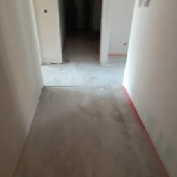 Usługi budowlane Piotr Skraburski - Korytarz w trakcie prac wykończeniowych: gładkie, białe ściany, wylewka podłogowa, widoczne ślady po taśmie malarskiej. Perspektywa z głębi pomieszczenia.