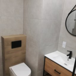 Usługi budowlane Piotr Skraburski - Nowoczesna łazienka z beżowymi płytkami, podwieszaną toaletą z drewnianą obudową i szafką pod umywalką z drewnianym frontem. Okrągłe lustro z czarną ramą.