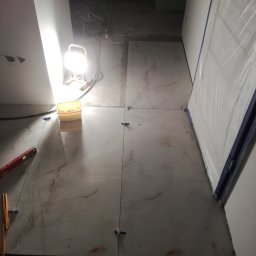 Usługi budowlane Piotr Skraburski - Układanie płytek podłogowych z użyciem krzyżyków dystansowych, oświetlone lampą budowlaną. Widoczny poziomica i narzędzia glazurnicze. Częściowo zakryte folią ochronną.