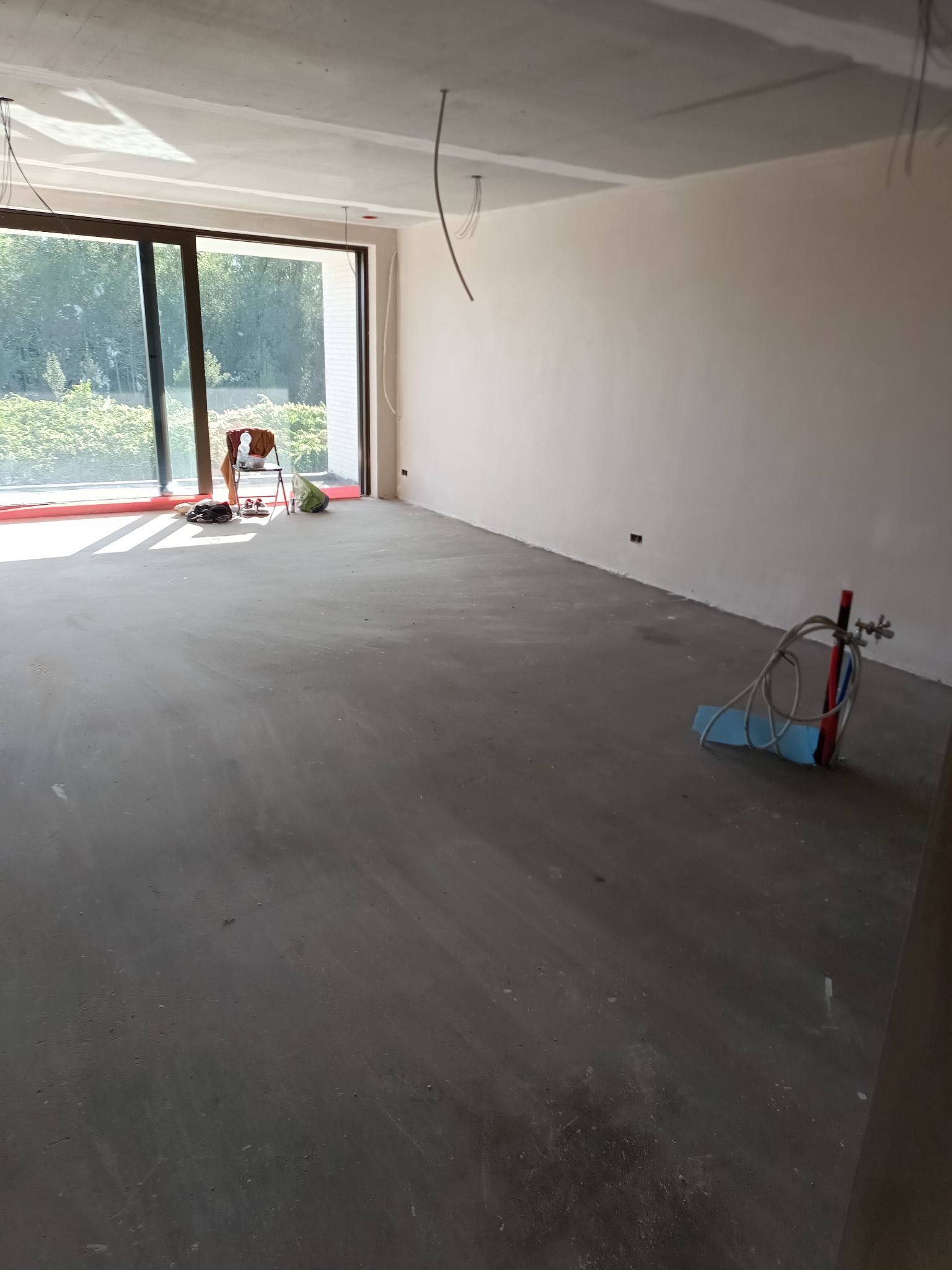 Surowe wnętrze w trakcie prac wykończeniowych: gładkie ściany, wylewka podłogowa, duże okno z widokiem na zieleń. Widoczne narzędzia i materiały budowlane.