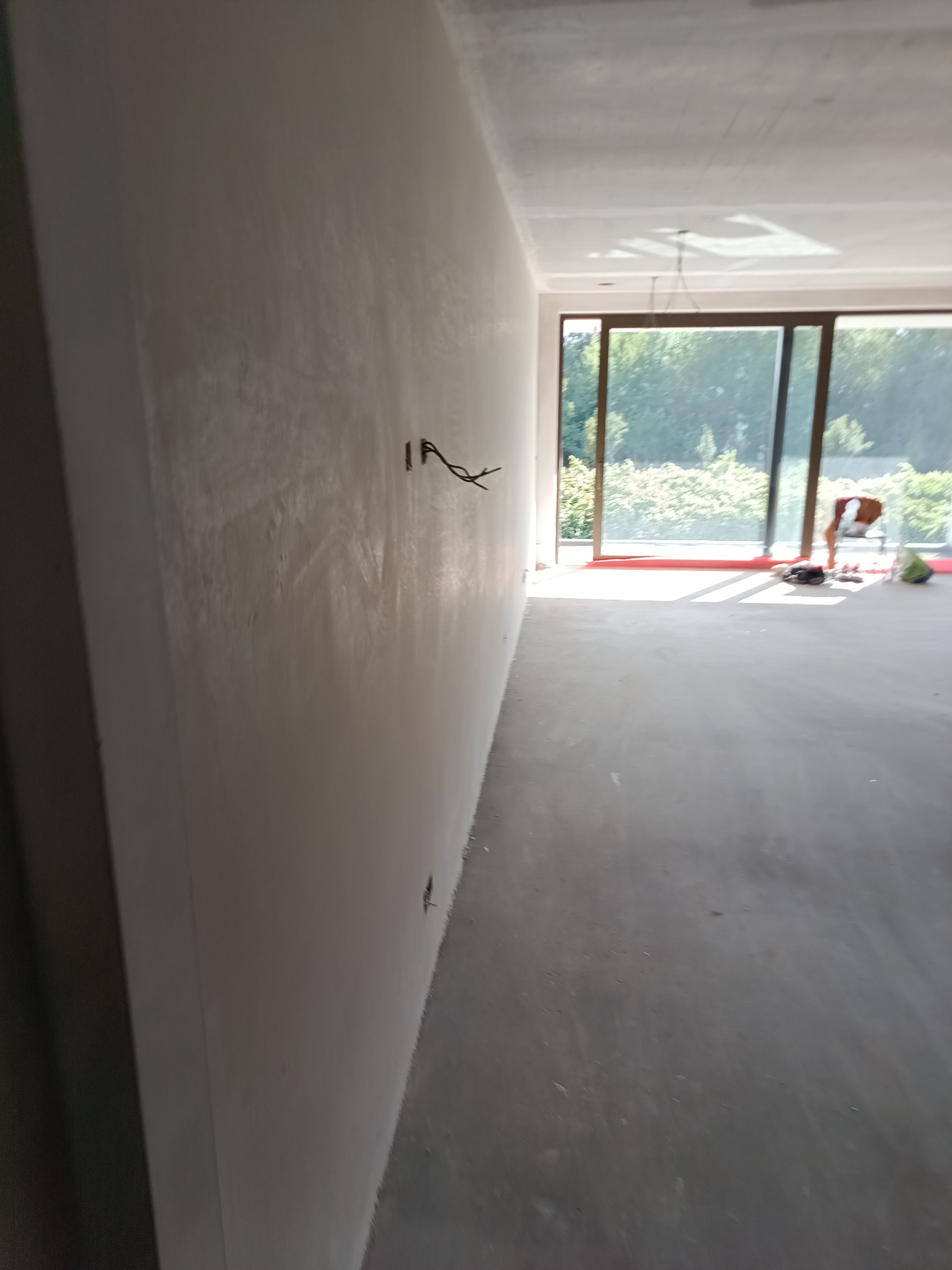 Ściana po szpachlowaniu z wystającymi kablami elektrycznymi. W tle widoczne okno balkonowe i osoba pracująca w jasnym, przestronnym wnętrzu.