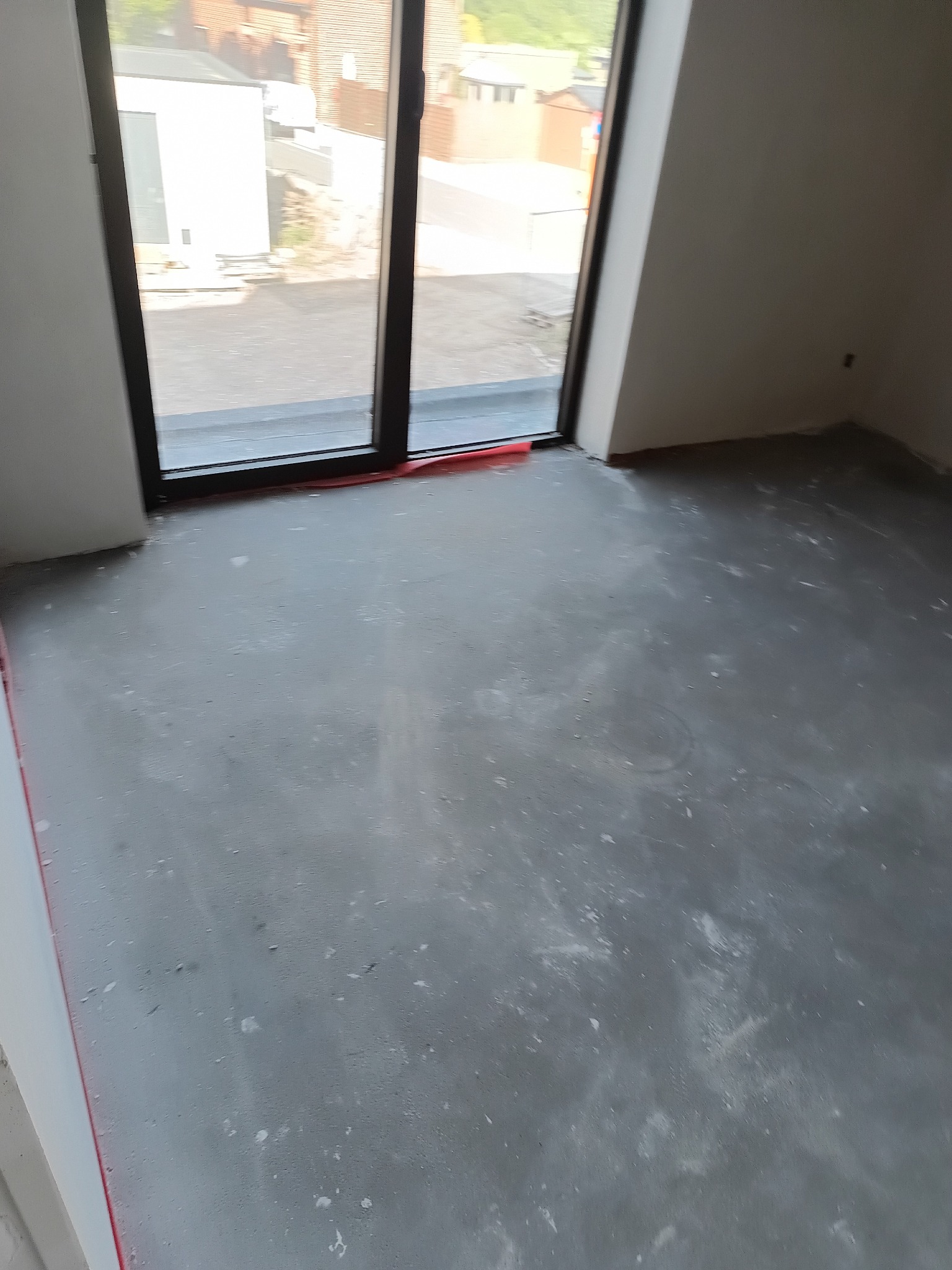 Wnętrze pokoju z wylewką samopoziomującą, przygotowane do dalszych prac wykończeniowych. Widoczne duże okno z czarną ramą i fragment budynku na zewnątrz.