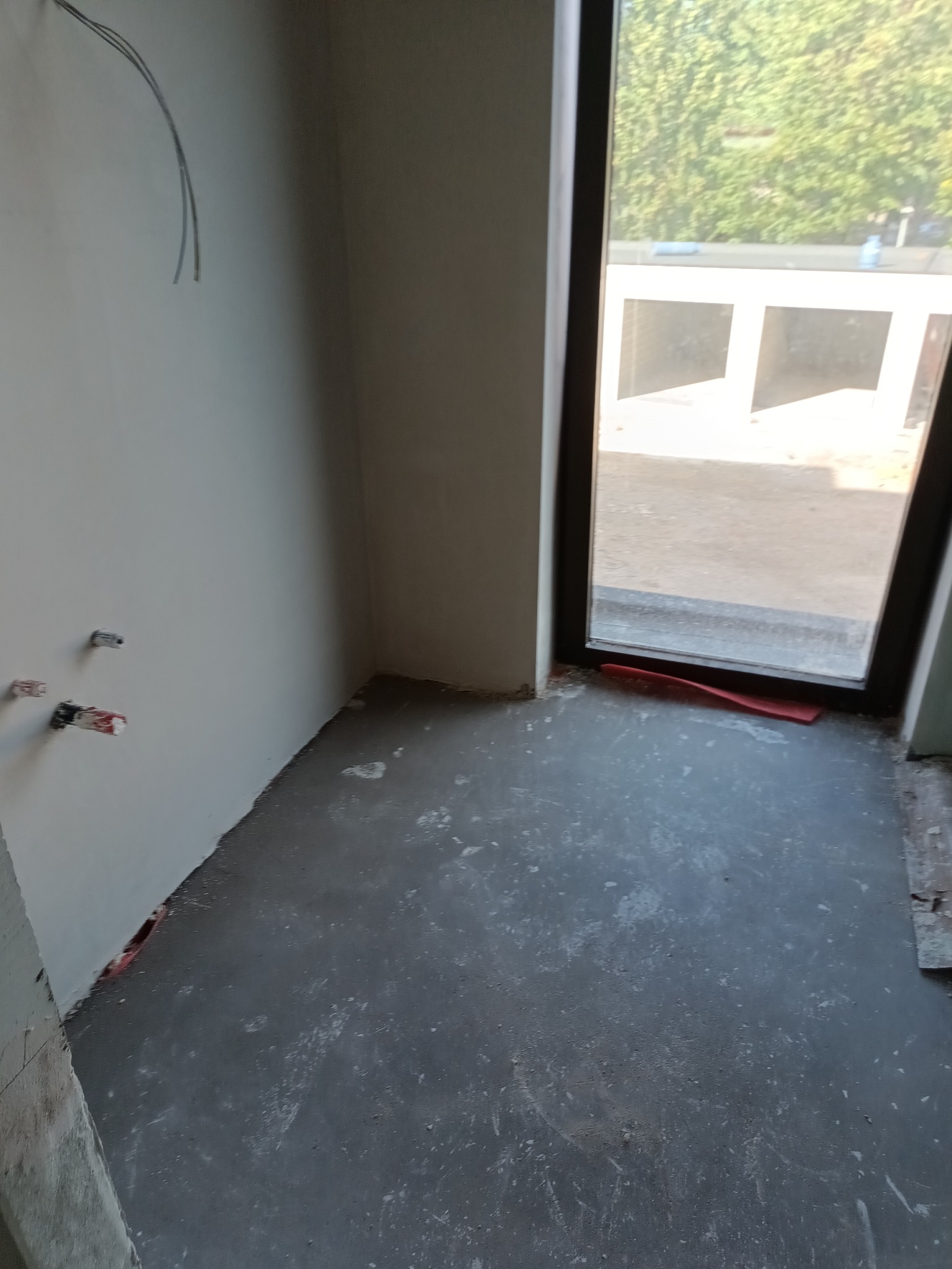 Surowe wnętrze w trakcie remontu: szpachlowane ściany, wylewka podłogowa, wystające kable i rury, okno z widokiem na zieleń. Etap przygotowania do dalszych prac wykończeniowych.