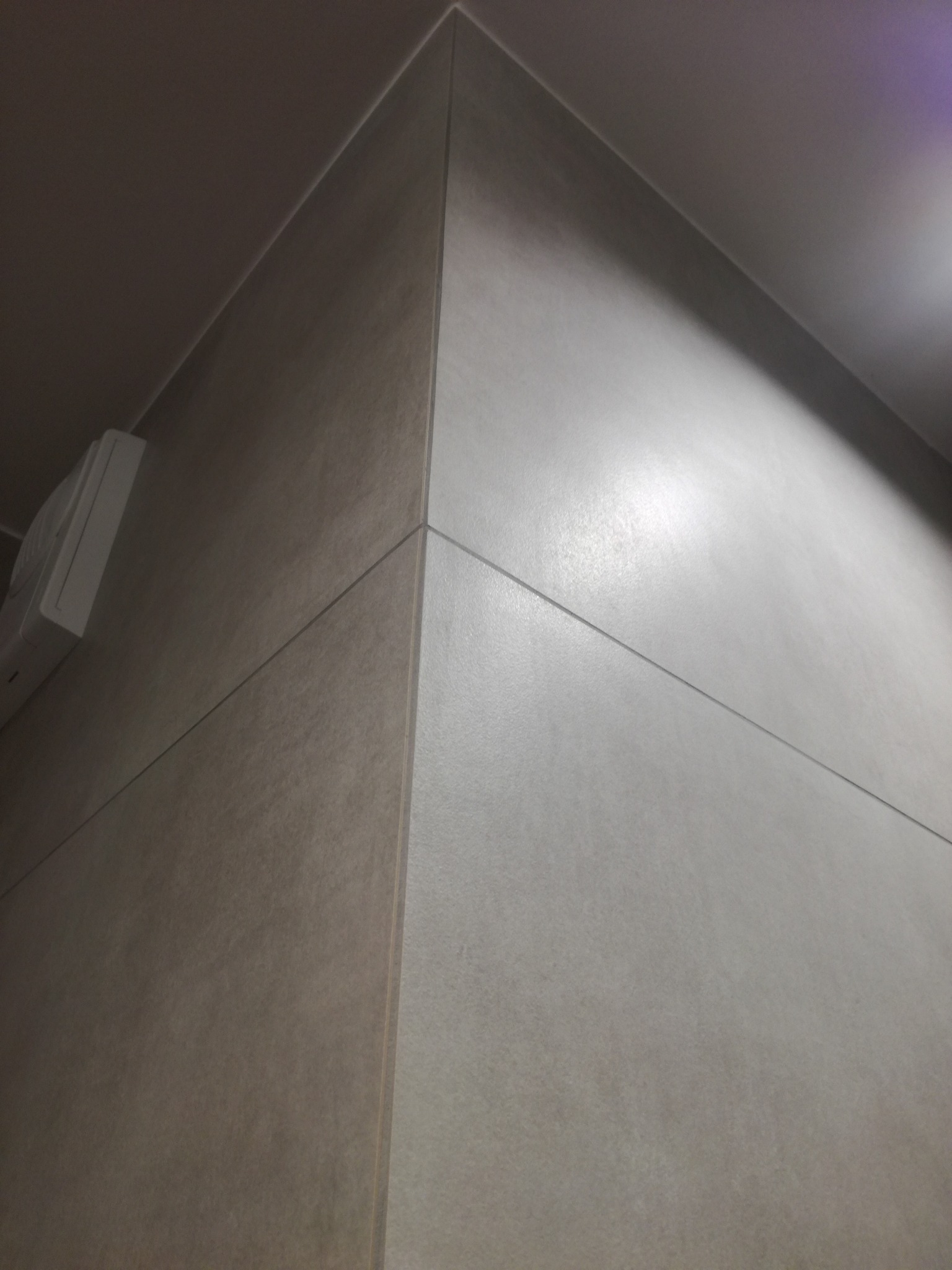 Szczegółowe ujęcie narożnika wykończonego beżowymi, matowymi płytkami ceramicznymi, z widocznymi fugami i elementem instalacji elektrycznej.