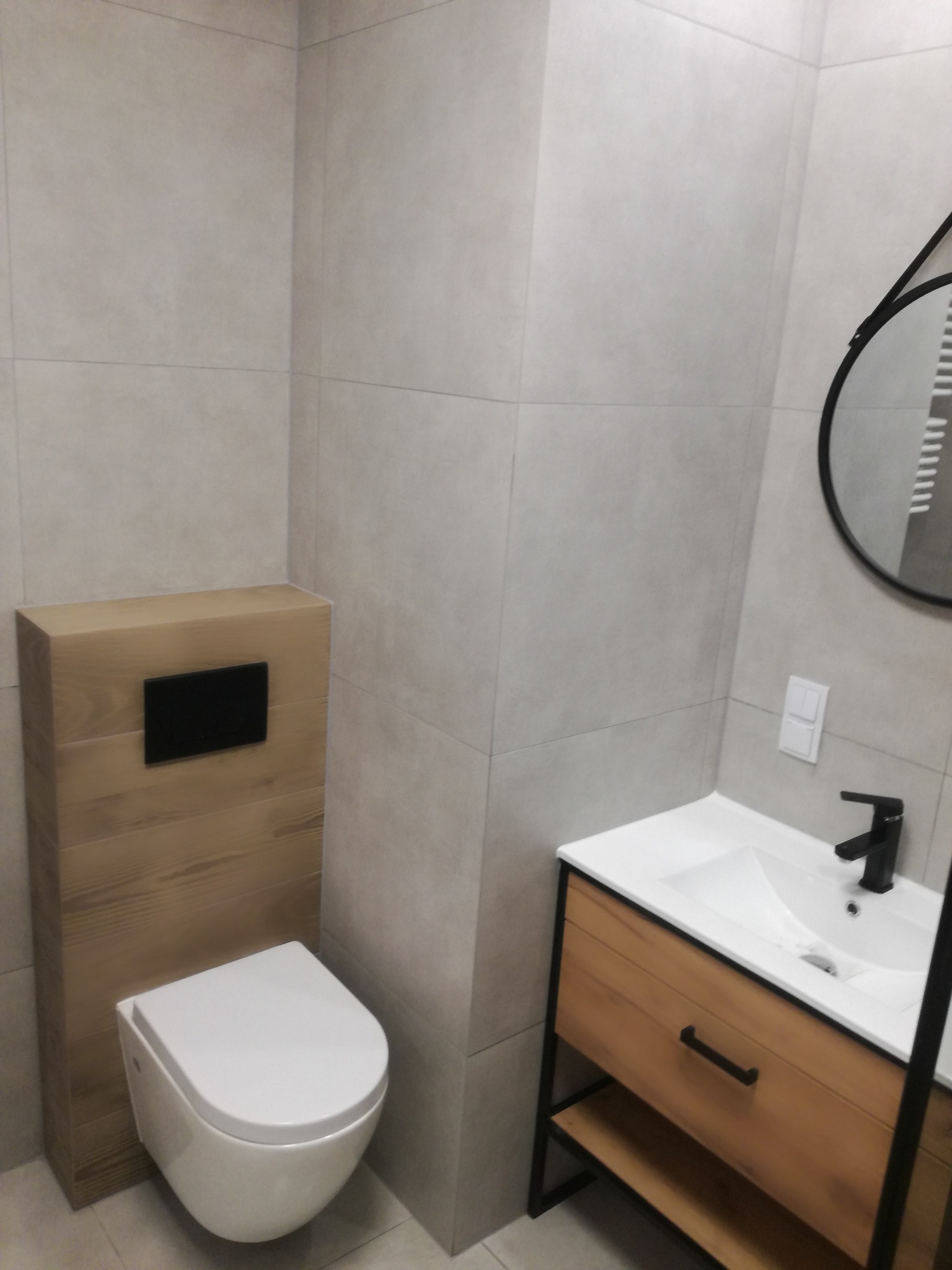 Nowoczesna łazienka z beżowymi płytkami, podwieszaną toaletą z drewnianą obudową i szafką pod umywalką z drewnianym frontem. Okrągłe lustro z czarną ramą.