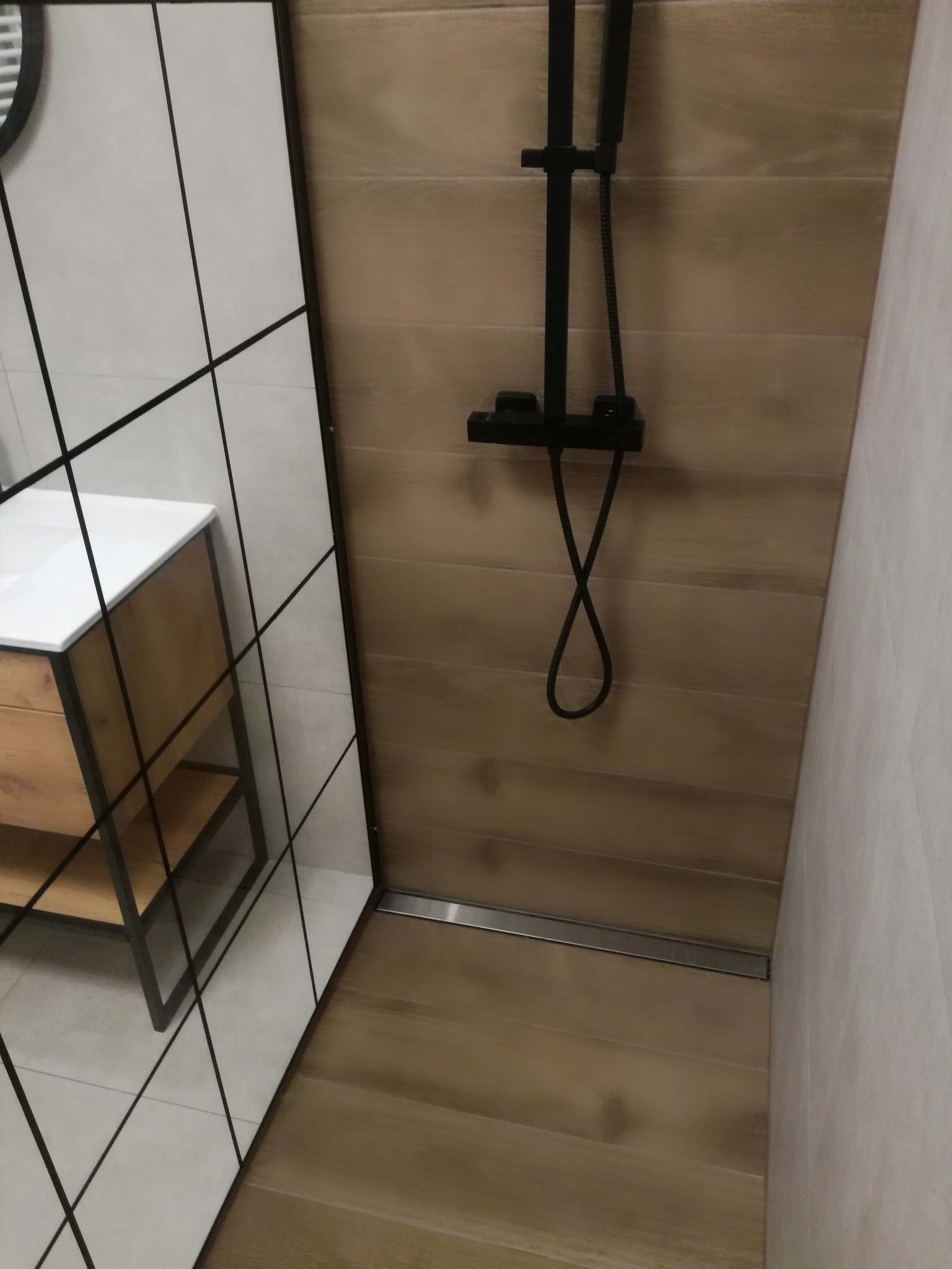 Nowoczesna łazienka z kabiną typu walk-in, wykończona płytkami imitującymi drewno i armaturą w kolorze czarnym. Widoczna umywalka z szafką w stylu loft.