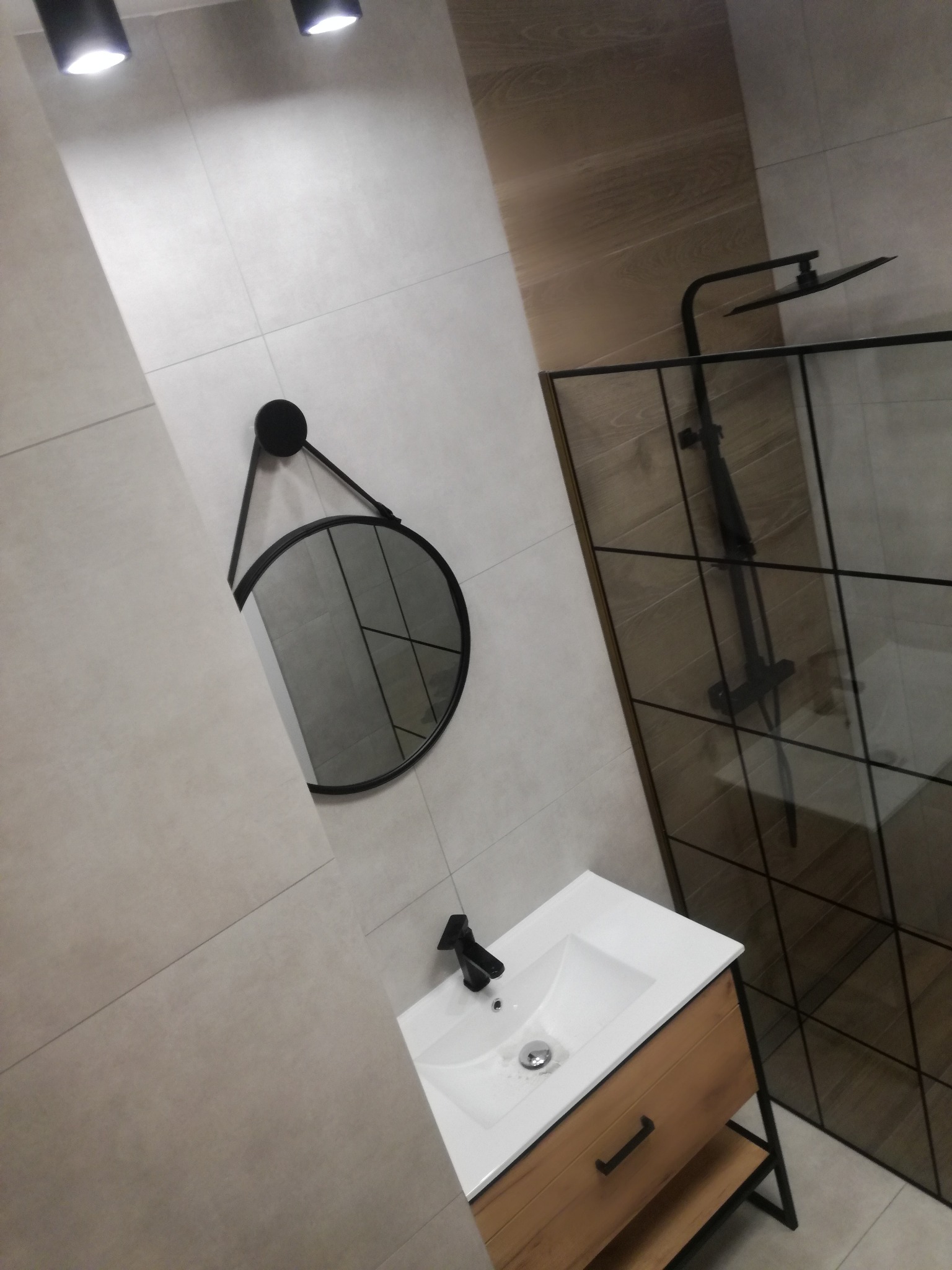 Nowoczesna łazienka z kabiną prysznicową typu walk-in, okrągłym lustrem i drewnianą szafką pod umywalką. Minimalistyczny design, czarne akcenty, jasne płytki.