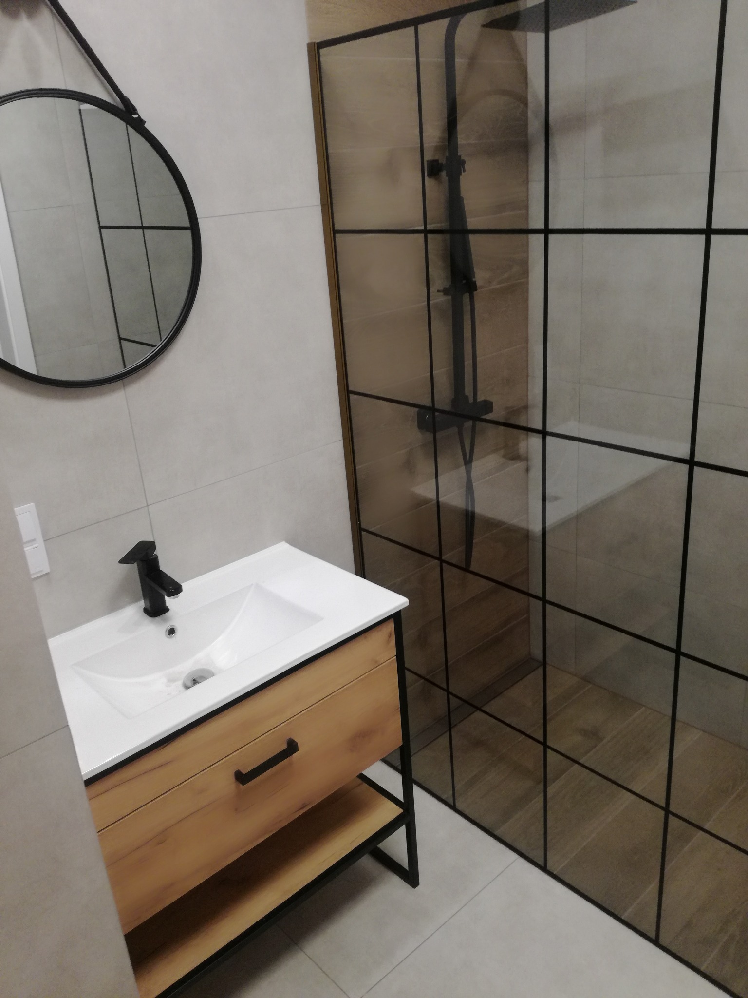 Nowoczesna łazienka: biała umywalka z drewnianą szafką, okrągłe lustro na szarej ścianie z płytek i kabina prysznicowa z czarnymi profilami.