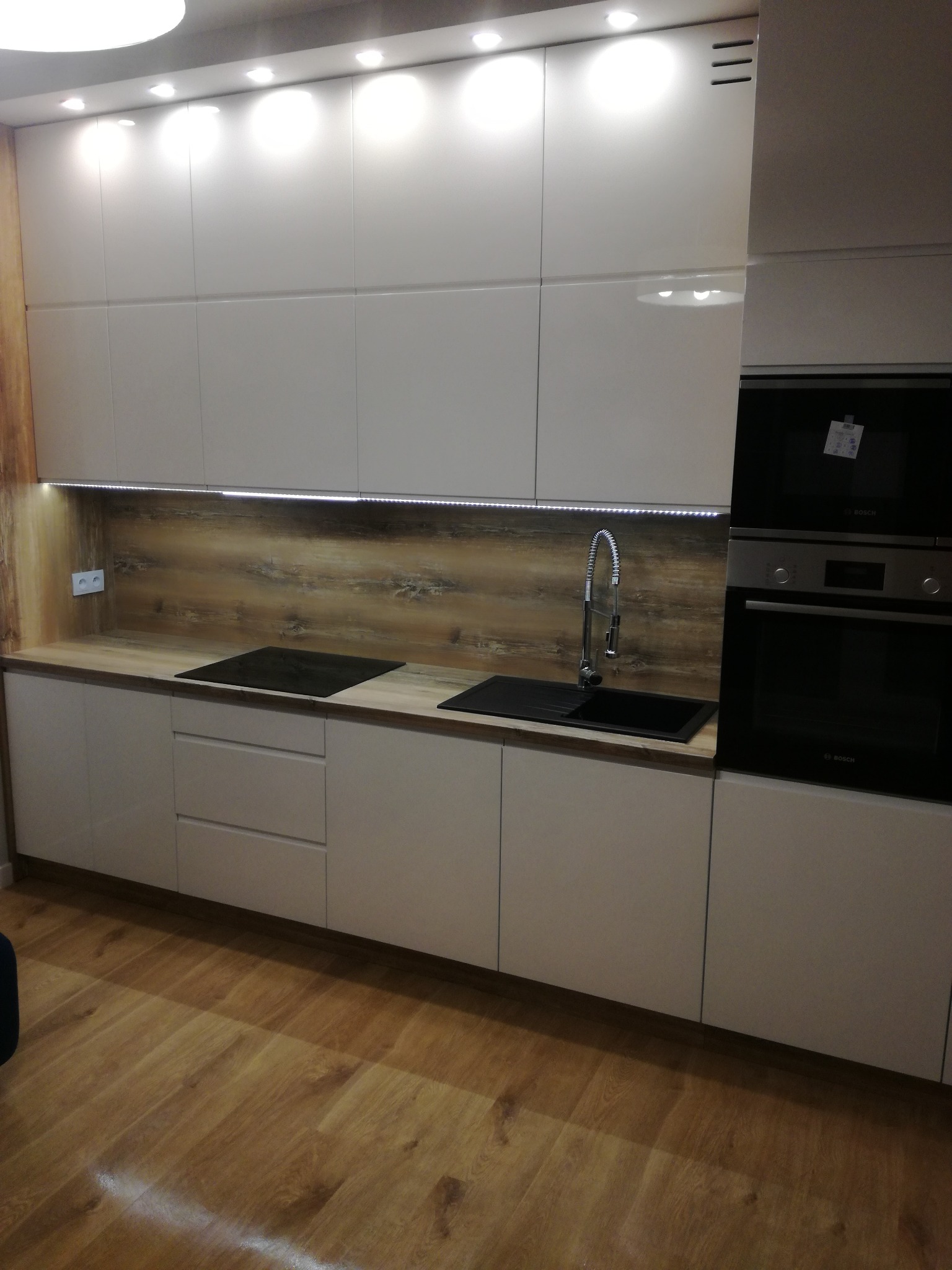 Nowoczesna kuchnia z białymi szafkami, drewnianym blatem i oświetleniem LED. Widoczny zlew, płyta indukcyjna i piekarnik. Parkiet na podłodze.