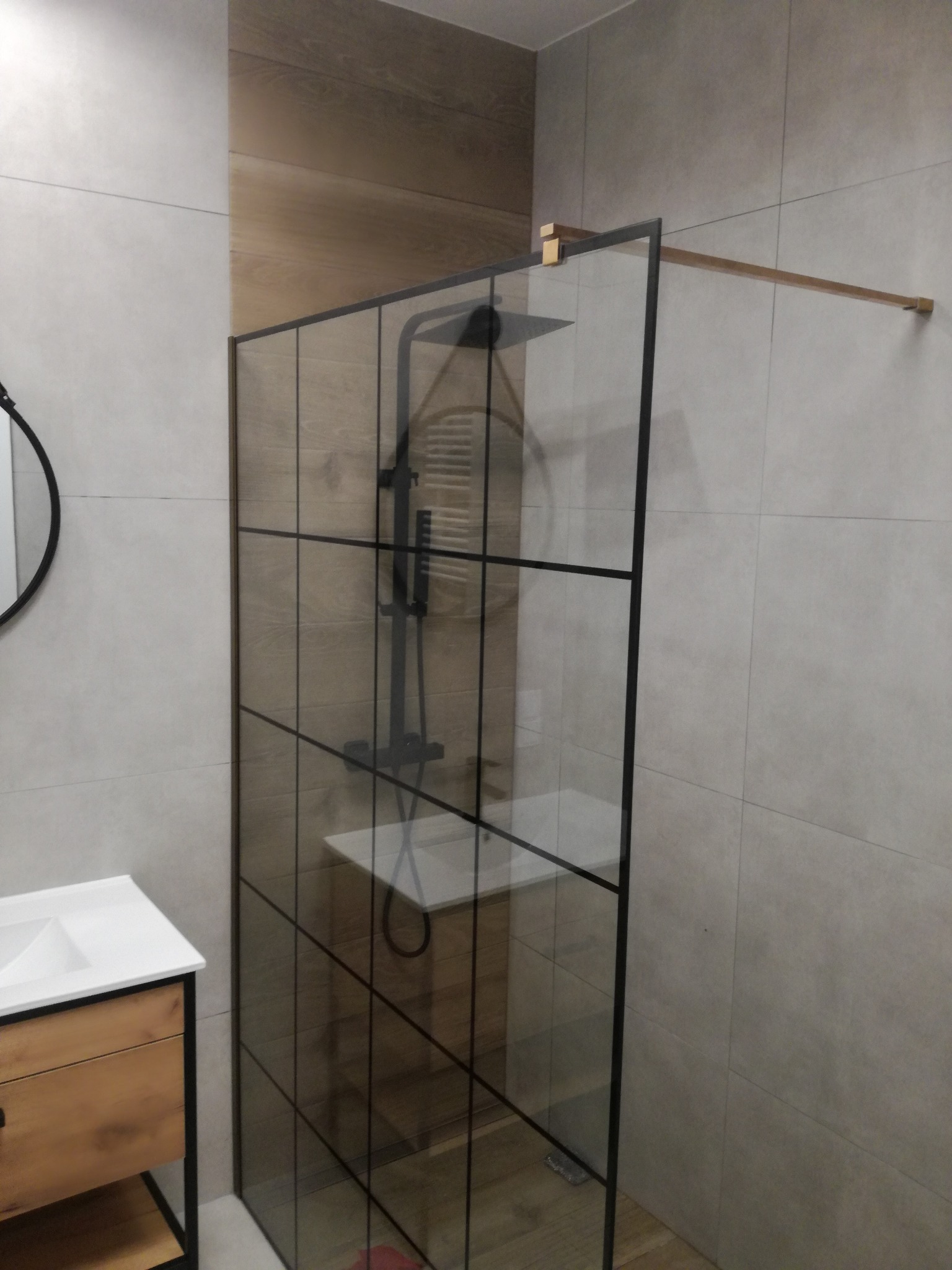 Nowoczesna kabina prysznicowa z czarnymi profilami i deszczownicą w wykończonej łazience. Widoczna umywalka z szafką z drewnianym frontem i szare płytki.