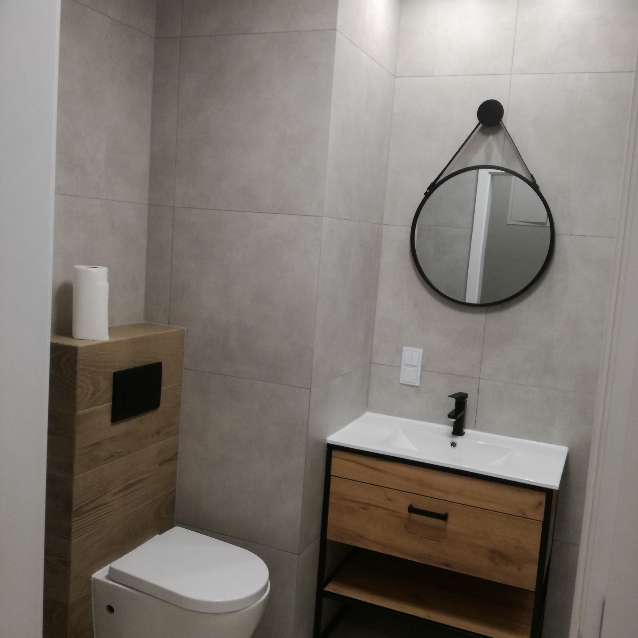 Wykończona łazienka z szarymi płytkami, drewnianą zabudową toalety i szafką pod umywalkę z okrągłym lustrem. Nowoczesny design.