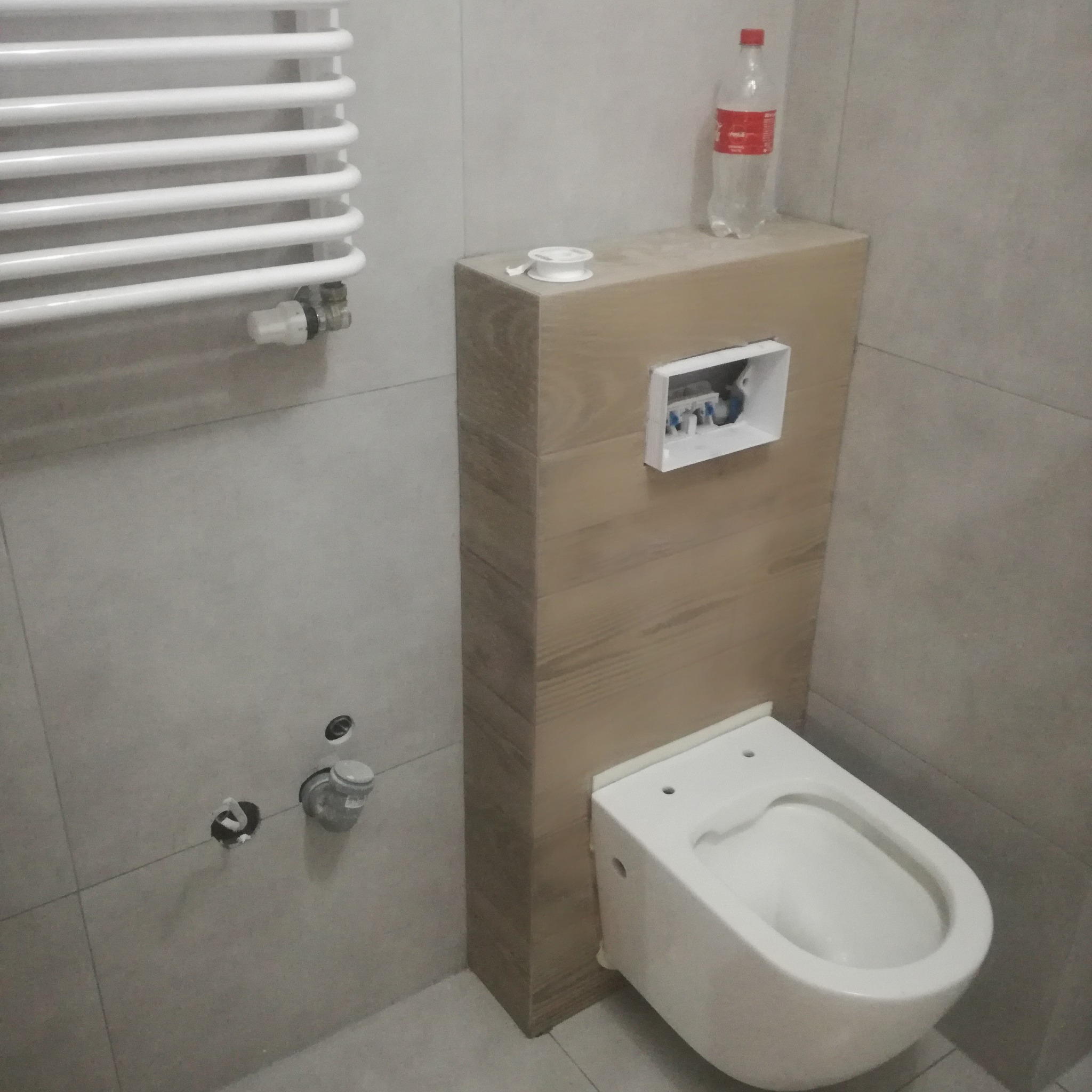 Wisząca toaleta w zabudowie z drewnopodobnych paneli, biały grzejnik drabinkowy na szarej, kafelkowanej ścianie. Widoczne przyłącza wodne i butelka na obudowie.