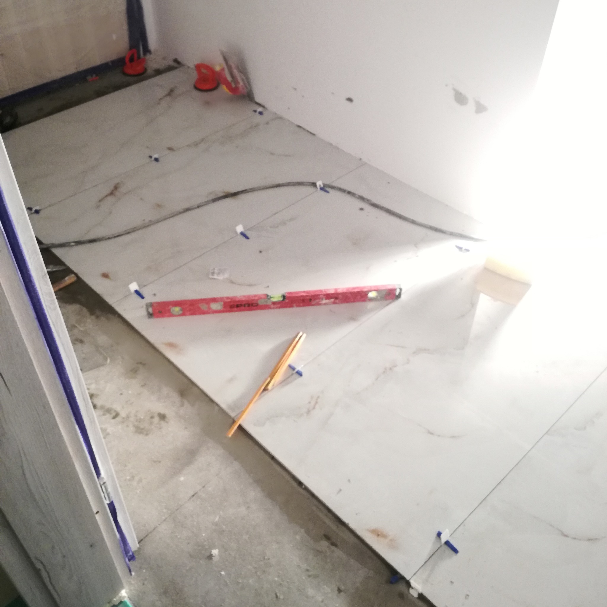 Układanie płytek podłogowych w łazience w Turku. Widoczny poziomica, ołówek i system poziomowania płytek. Białe płytki imitujące marmur.