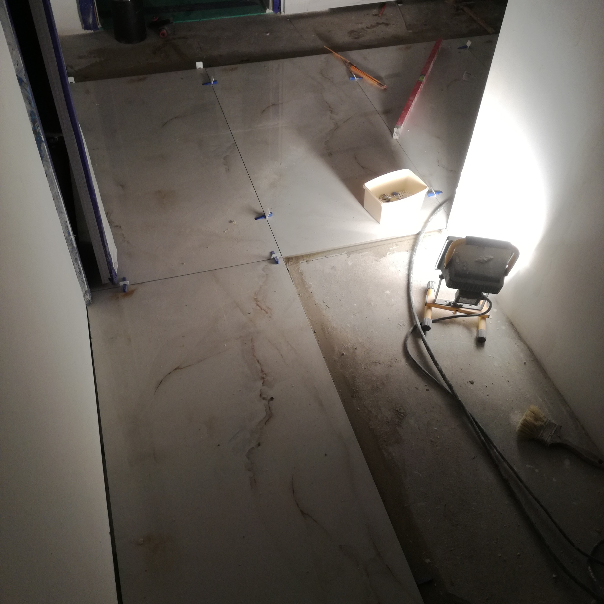 Układanie płytek podłogowych z użyciem krzyżyków dystansowych, oświetlone lampą budowlaną. Widoczny poziomica i pojemnik z akcesoriami.