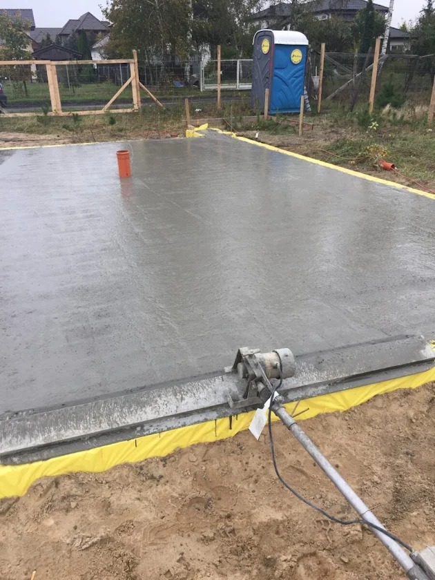 Wyrównywanie wylewki betonowej na fundamencie z użyciem listwy wibracyjnej. Widoczna folia izolacyjna i przenośna toaleta w tle.