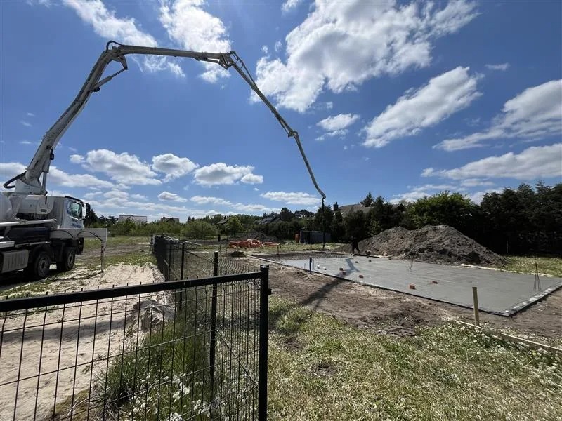 Betonowanie płyty fundamentowej z użyciem pompy do betonu na działce budowlanej w słoneczny dzień. Widoczne zbrojenie i przygotowany teren.