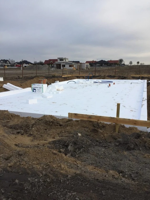 Ocieplone fundamenty pod budowę domu jednorodzinnego. Widoczne białe płyty styropianowe, drewniane szalunki i instalacje. W tle zabudowa jednorodzinna.