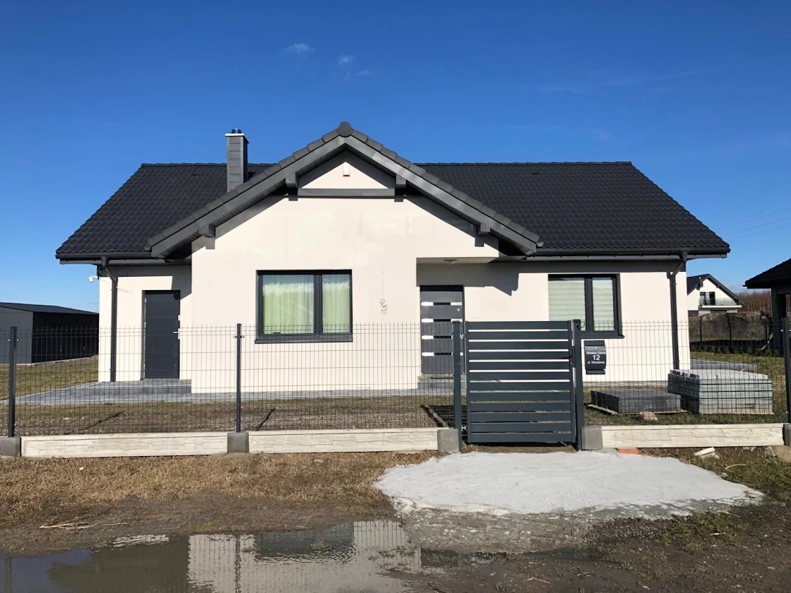 Nowoczesny dom jednorodzinny z ciemną dachówką i szarą stolarką okienną, ogrodzony siatką panelową na podmurówce. Widoczne wejście i brama wjazdowa.