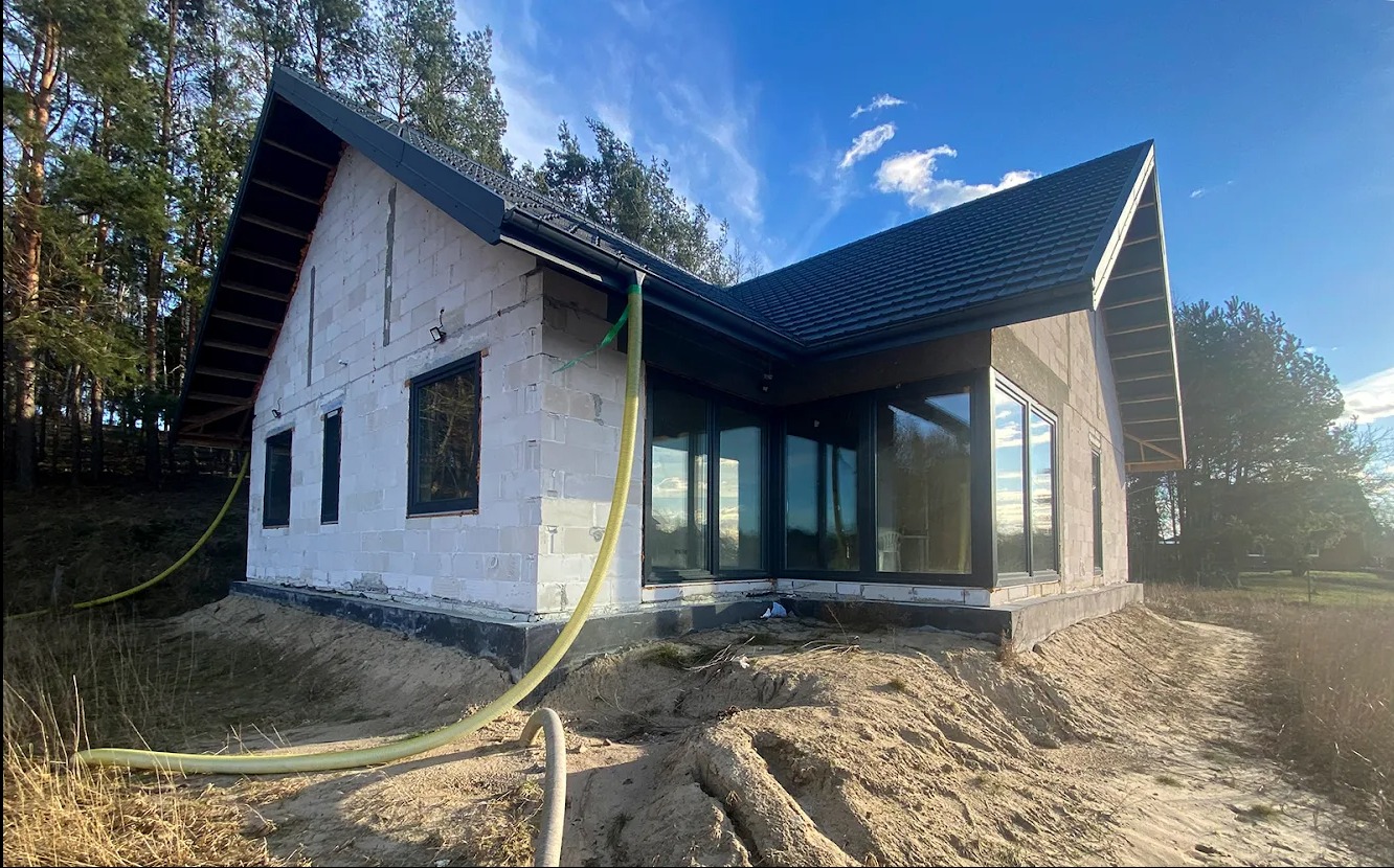 Niedokończony dom z białego pustaka z czarną stolarką okienną i dachem. Żółty wąż na piasku. Budowa w lesie, błękitne niebo.