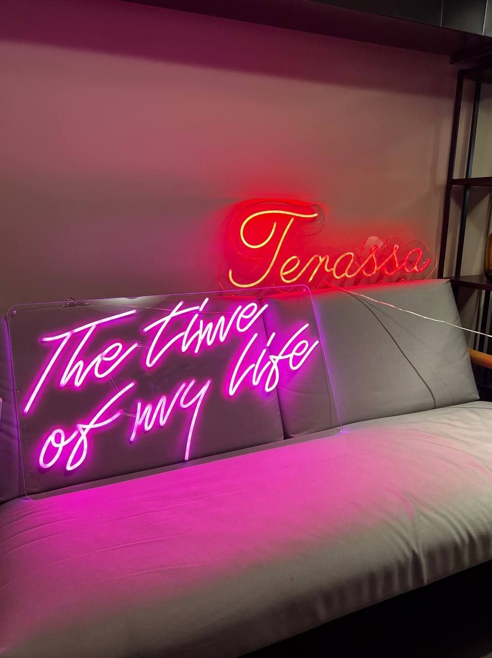 Dwa neony: różowy z napisem 'The time of my life' na pleksi i czerwono-żółty 'Terassa', leżą na szarej sofie. Ujęcie z bliska, widoczny fragment regału.