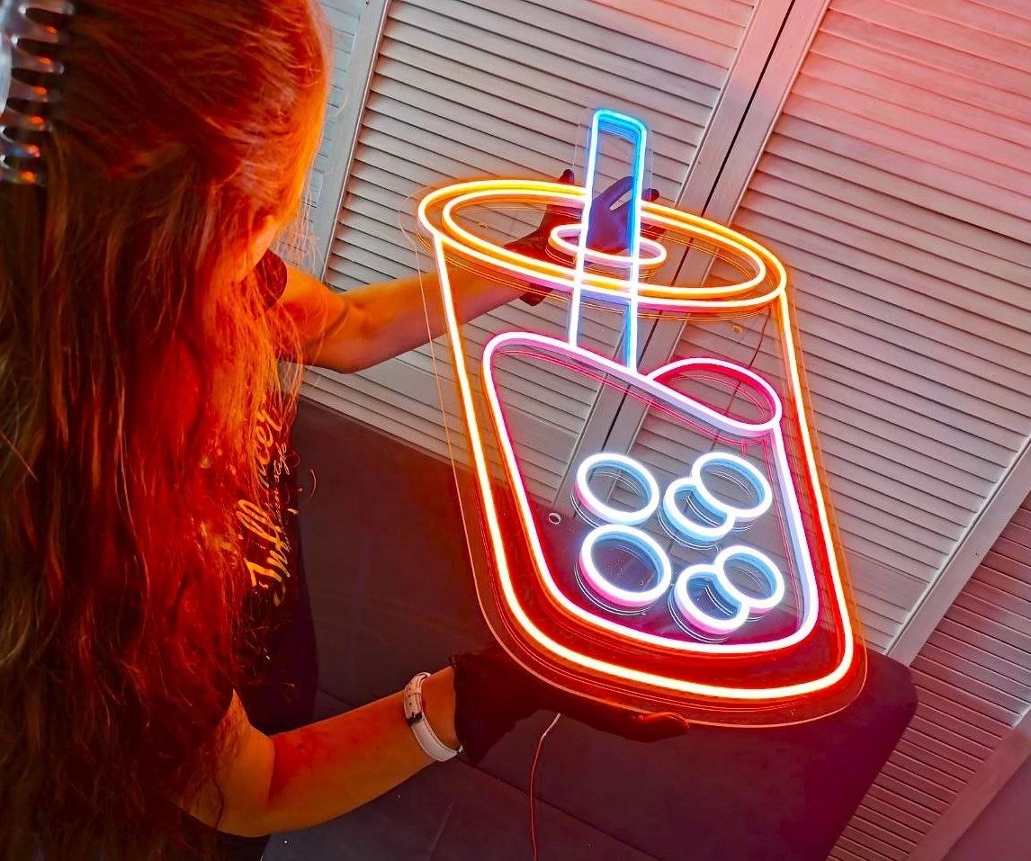 Neonowy szyld w kształcie kubka Bubble Tea, trzymany przez osobę. Intensywne kolory neonu: pomarańczowy, różowy i niebieski. Białe, drewniane panele w tle.