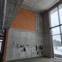 DOMI-TEAM DOMINIKA BURACZEWSKA USŁUGI TYNKARSKIE - Surowy stan wnętrza: ściany z betonu architektonicznego, fragment muru z cegły dziurawki, instalacje pod sufitem, duże okno z widokiem na zimowy krajobraz.