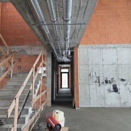 DOMI-TEAM DOMINIKA BURACZEWSKA USŁUGI TYNKARSKIE - Surowy stan budynku w trakcie prac: widoczne ceglane ściany, betonowy strop z instalacjami, schody z drewnianą barierką i nagrzewnica na podłodze.