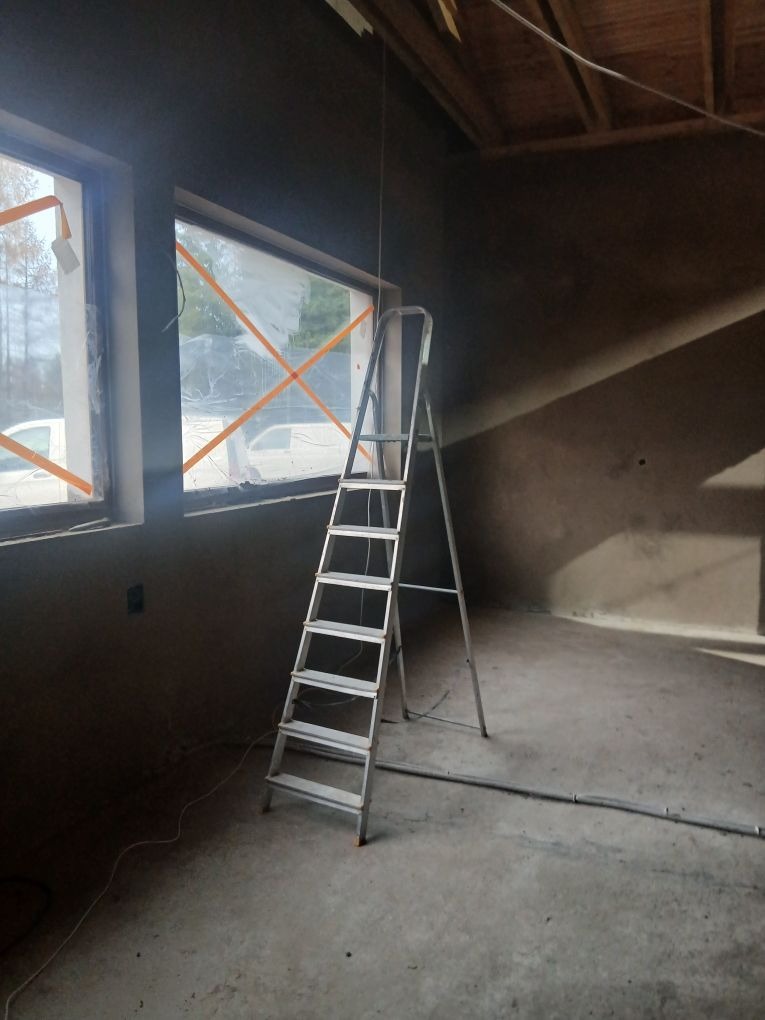 Wnętrze w trakcie prac. Ściany pokryte świeżym, ciemnym tynkiem, okna zabezpieczone folią. Aluminiowa drabina oparta o ścianę. Widoczne przewody na podłodze.