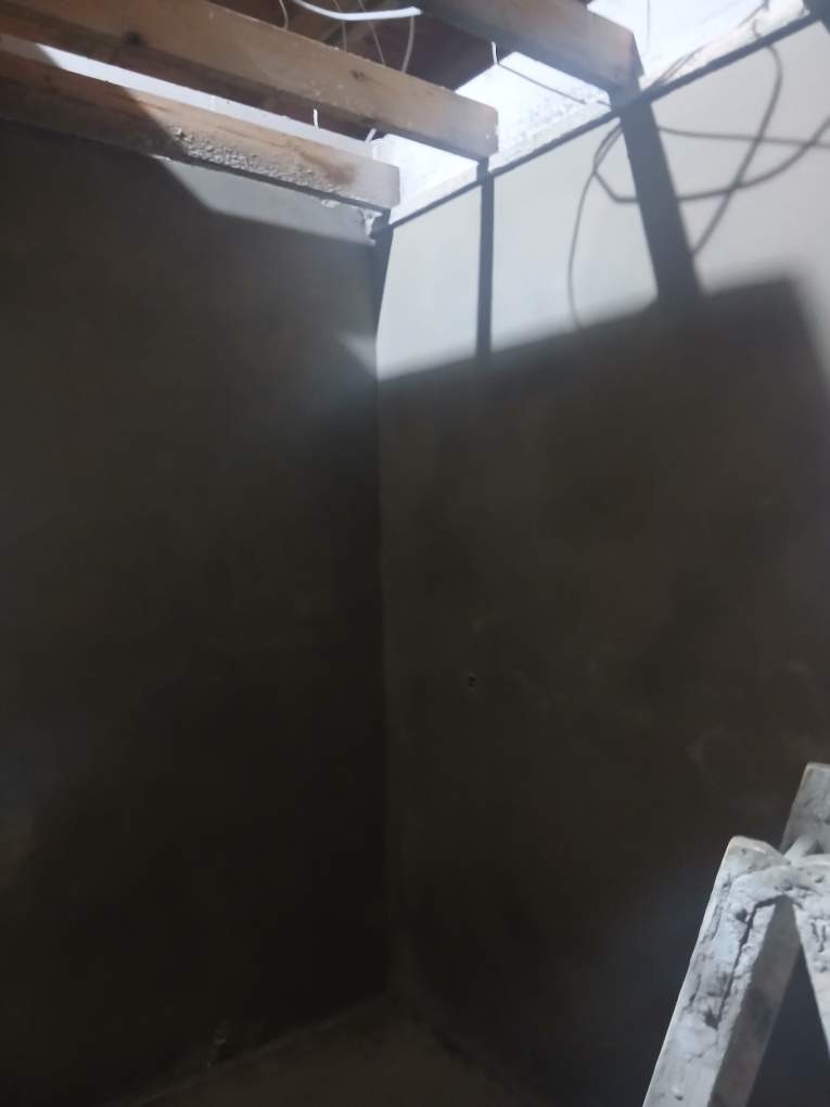 Świeżo otynkowane ściany w rogu pomieszczenia, widoczne drewniane elementy konstrukcyjne sufitu i fragment drabiny. Szare, gładkie powierzchnie ścian po tynkowaniu.