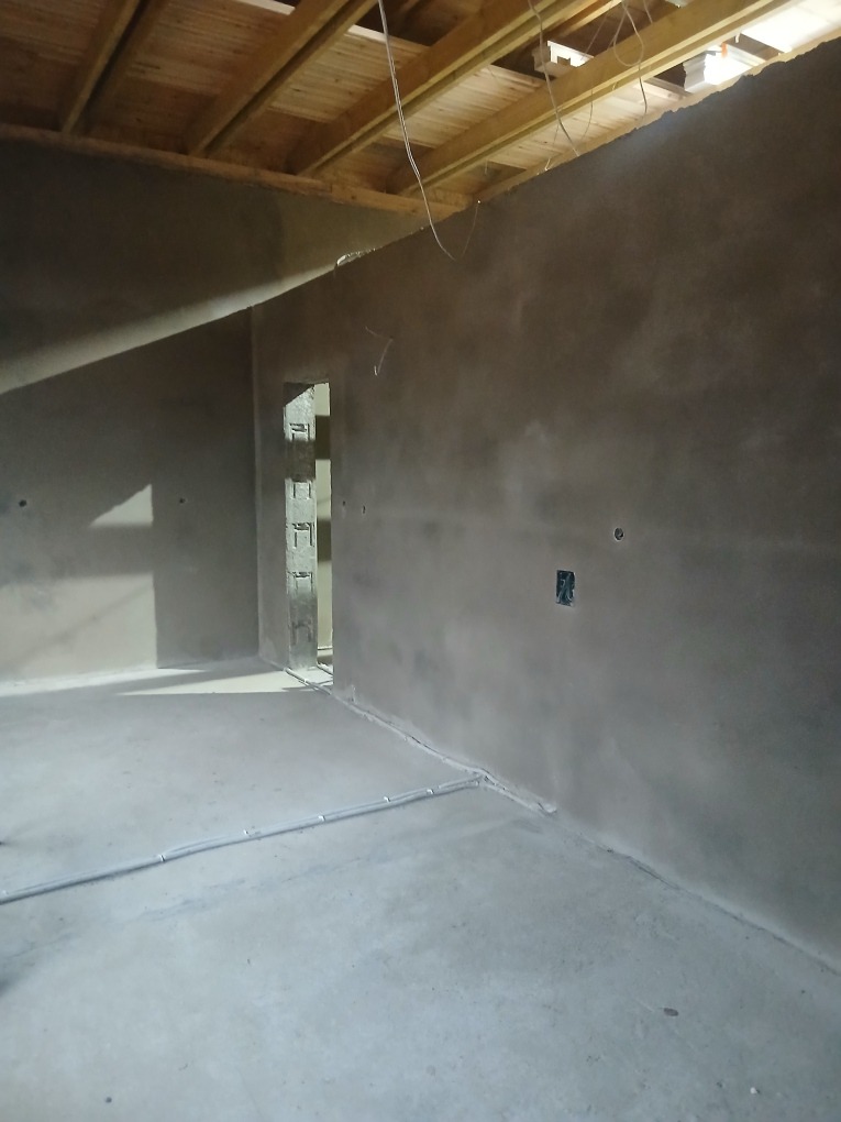 Wnętrze po nałożeniu tynku: szare ściany, surowy drewniany strop, widoczne przewody elektryczne w ścianie i na podłodze. Surowy stan wykończenia.