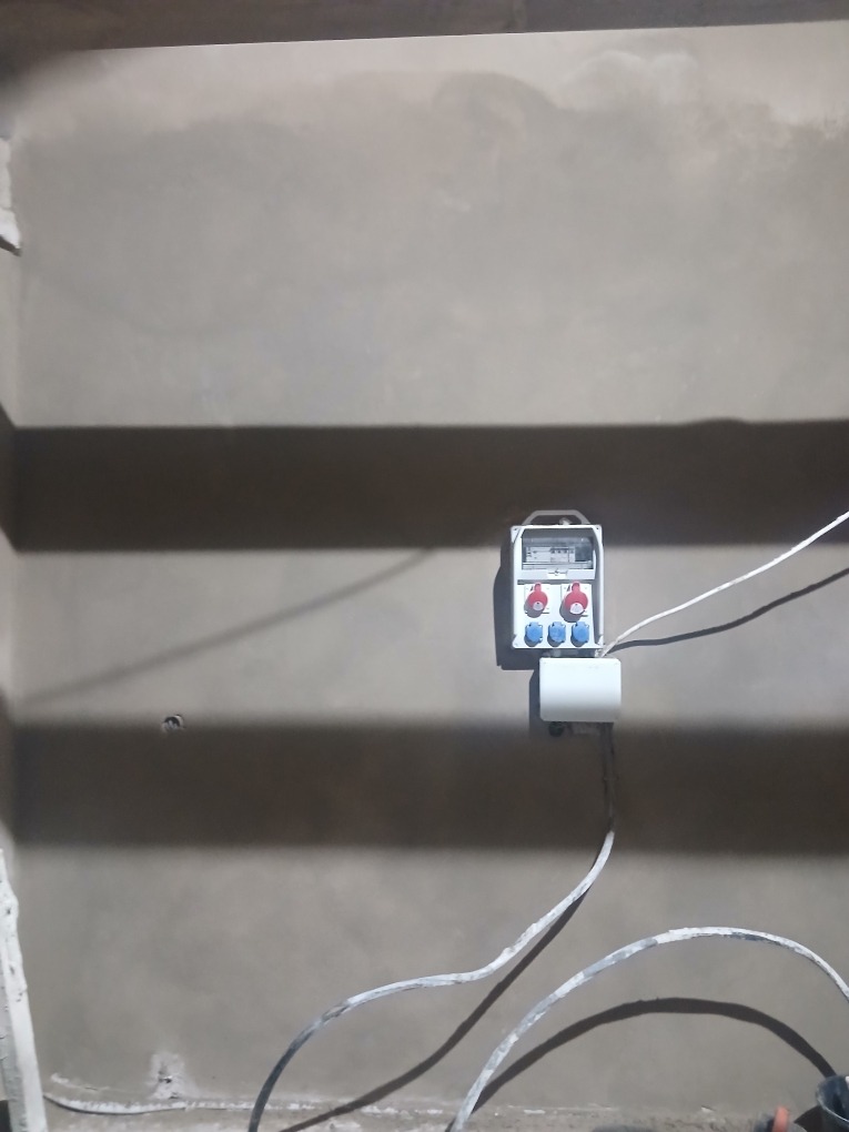 Świeżo nałożony, szary tynk na ścianie wewnętrznej, z widocznym gniazdkiem elektrycznym i przewodami. Nierównomierna faktura tynku w górnej części ściany.