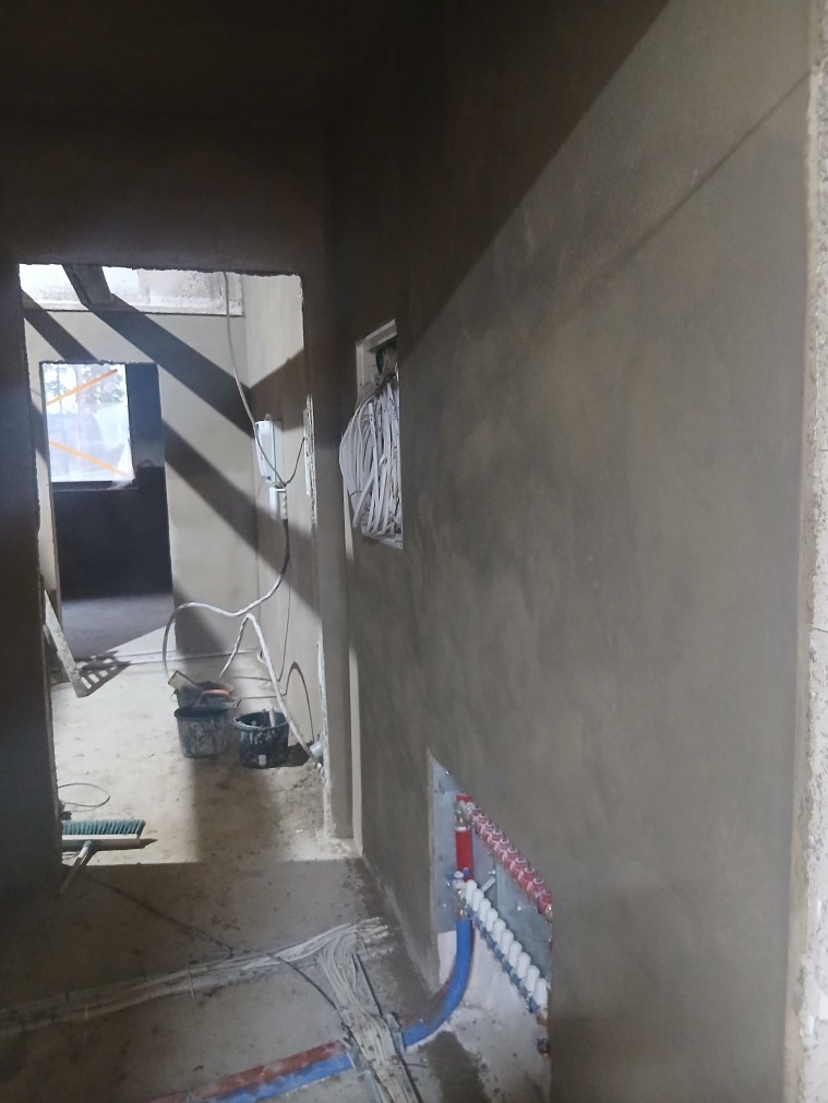 Świeżo otynkowane ściany w korytarzu, widoczne instalacje elektryczne i ogrzewania podłogowego. Surowy stan wykończenia wnętrza, przygotowane do dalszych prac.