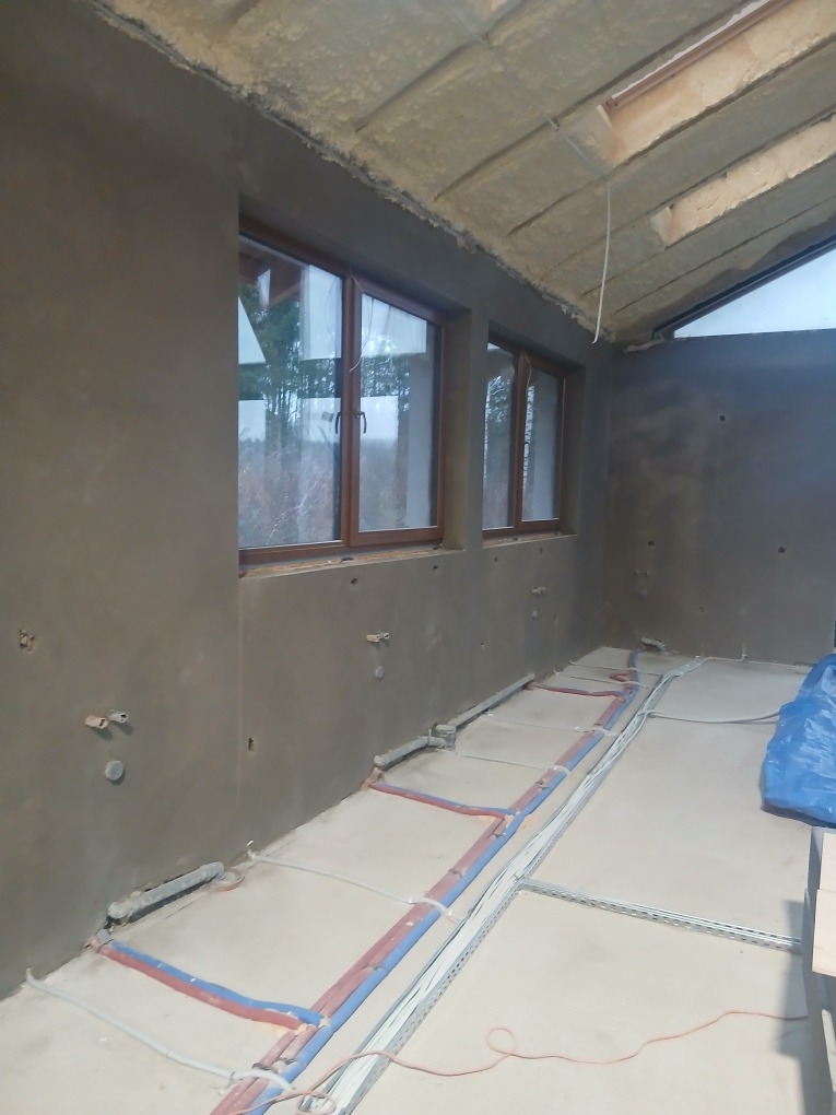 Wnętrze w trakcie prac wykończeniowych: ściany pokryte świeżym tynkiem, widoczne instalacje podłogowe, okna z drewnianymi ramami i sufit z izolacją.