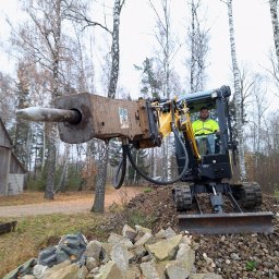 DARIUSZ CZYKWIN MERIDAN - Operator w żółtej kurtce obsługuje minikoparkę z młotem hydraulicznym, krusząc betonowe elementy na tle drzew i drewnianego budynku.