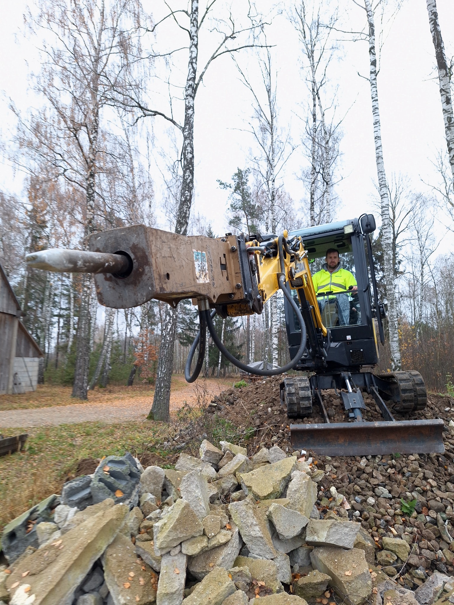 Operator w żółtej kurtce obsługuje minikoparkę z młotem hydraulicznym, krusząc betonowe elementy na tle drzew i drewnianego budynku.