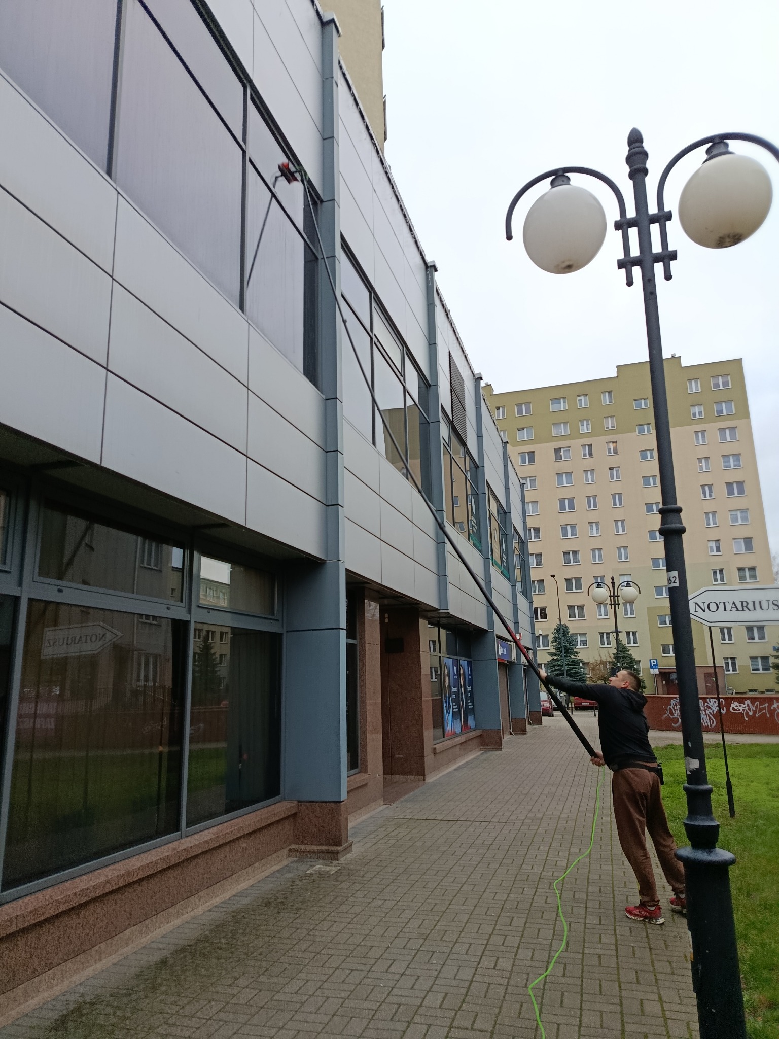 Mężczyzna myje okna na wysokości w biurowcu za pomocą specjalistycznego sprzętu. Widoczny budynek z szarymi panelami i duże okna. W tle budynek mieszkalny.