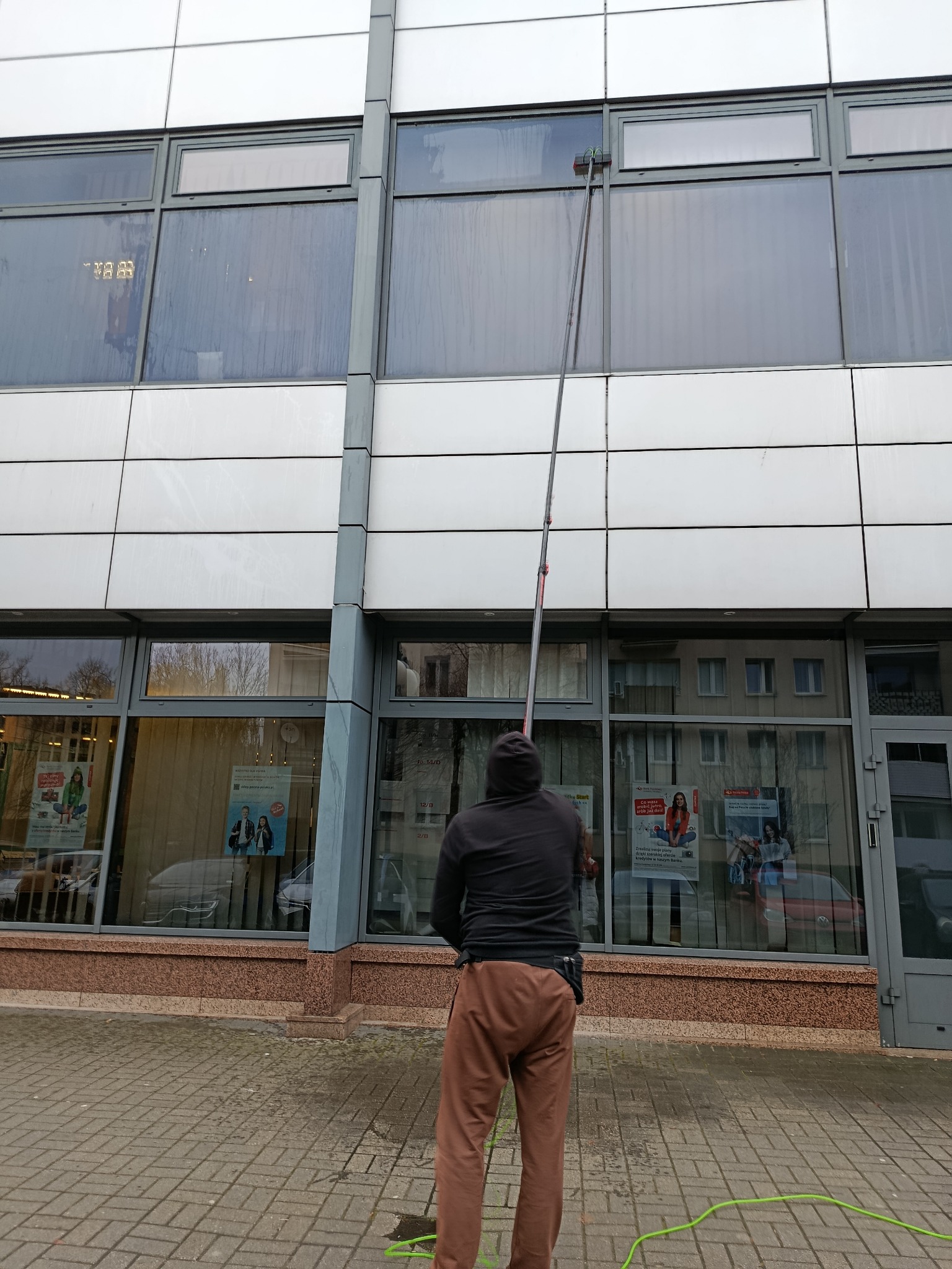 Mężczyzna w brązowych spodniach myje okna na wysokości w budynku biurowym za pomocą długiego kija z szczotką. Widoczne plakaty za szybami.