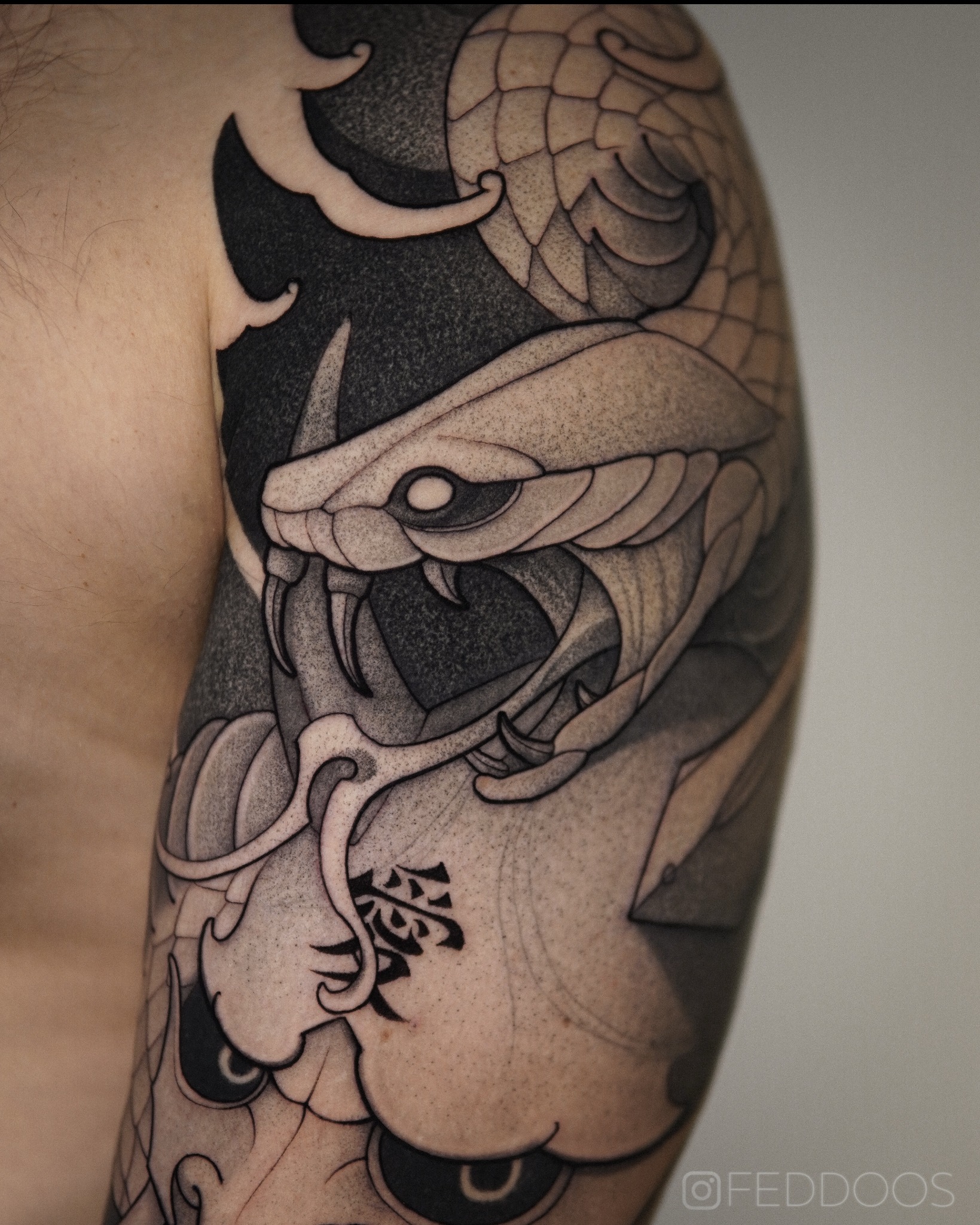 Ramię z czarno-białym tatuażem przedstawiającym głowę kobry z otwartą paszczą i widocznymi kłami, wzór w stylu japońskim z motywem fal i znakiem Kanji.